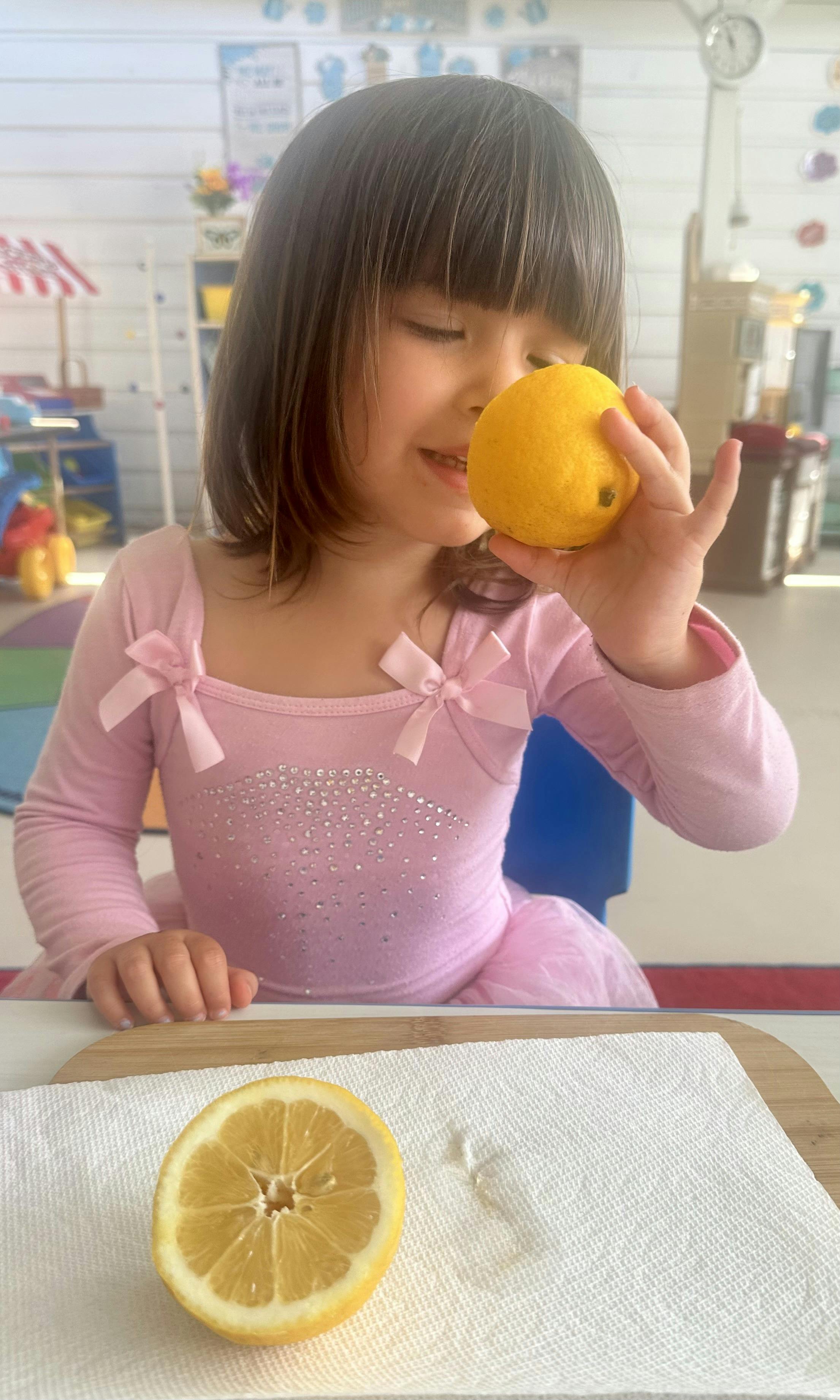 Child in a pink dress holding a lemon up to their nose in a classroom