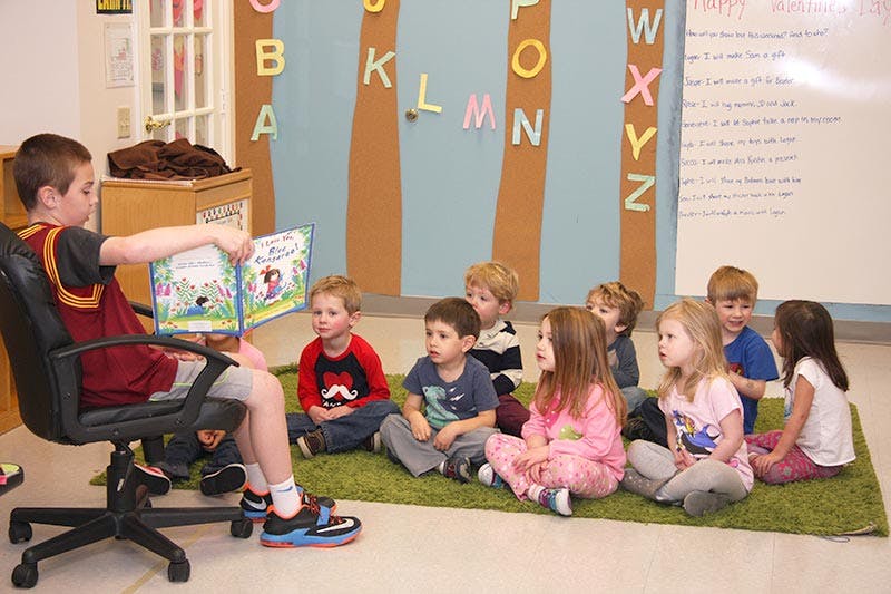 Child reading a book to a group of children sitting on a rug in a classroom