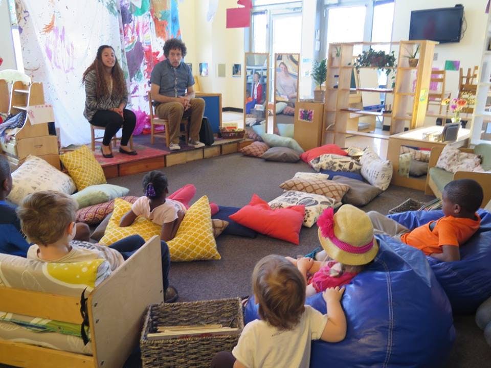 Two adults sitting on a stage in front of children sitting on pillows