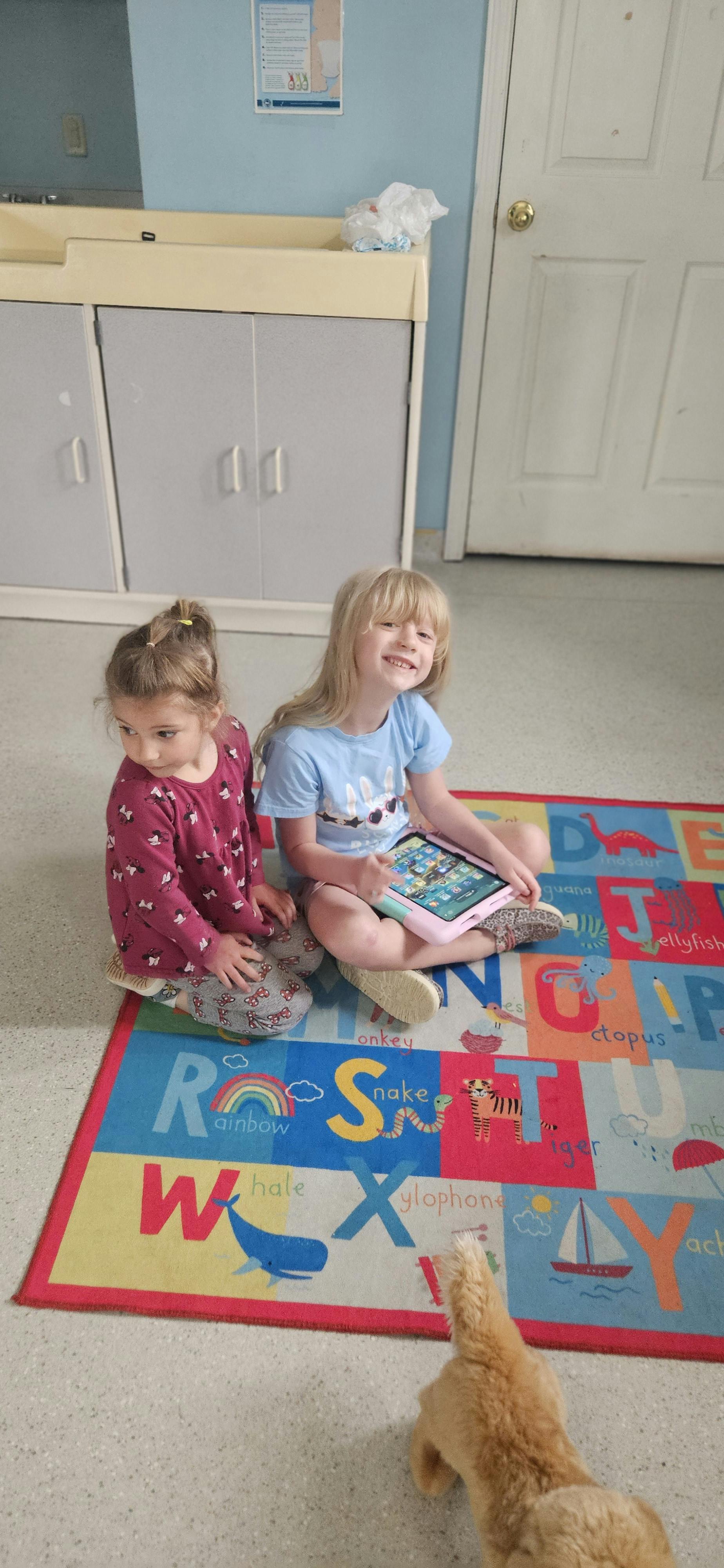 Two children sitting on an alphabet rug with a tablet