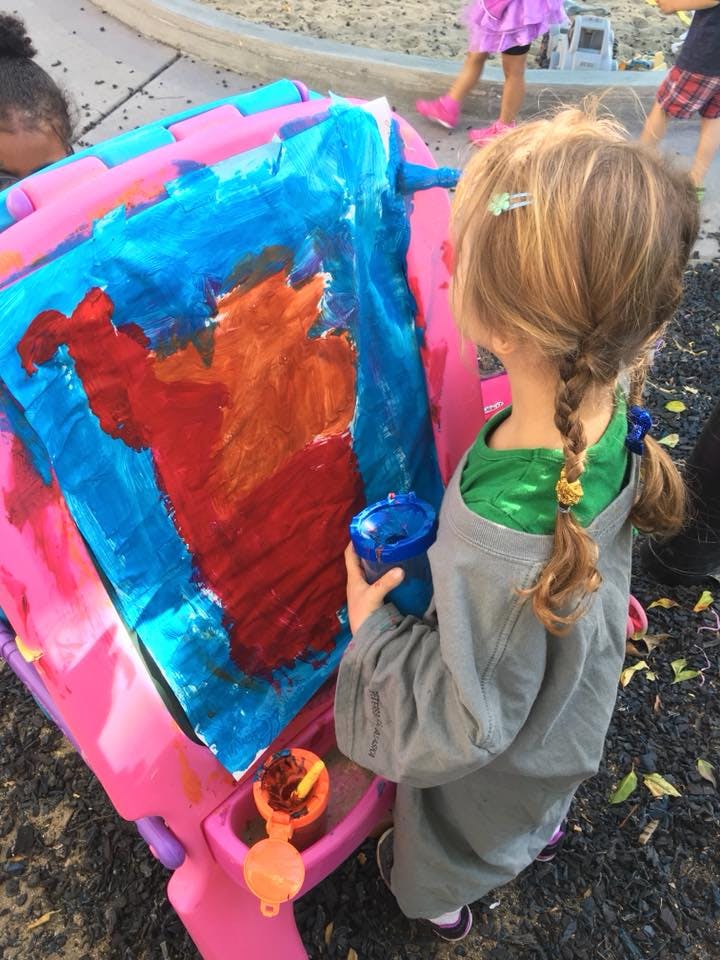 Child holding a cup next to a colorful painting on an easel