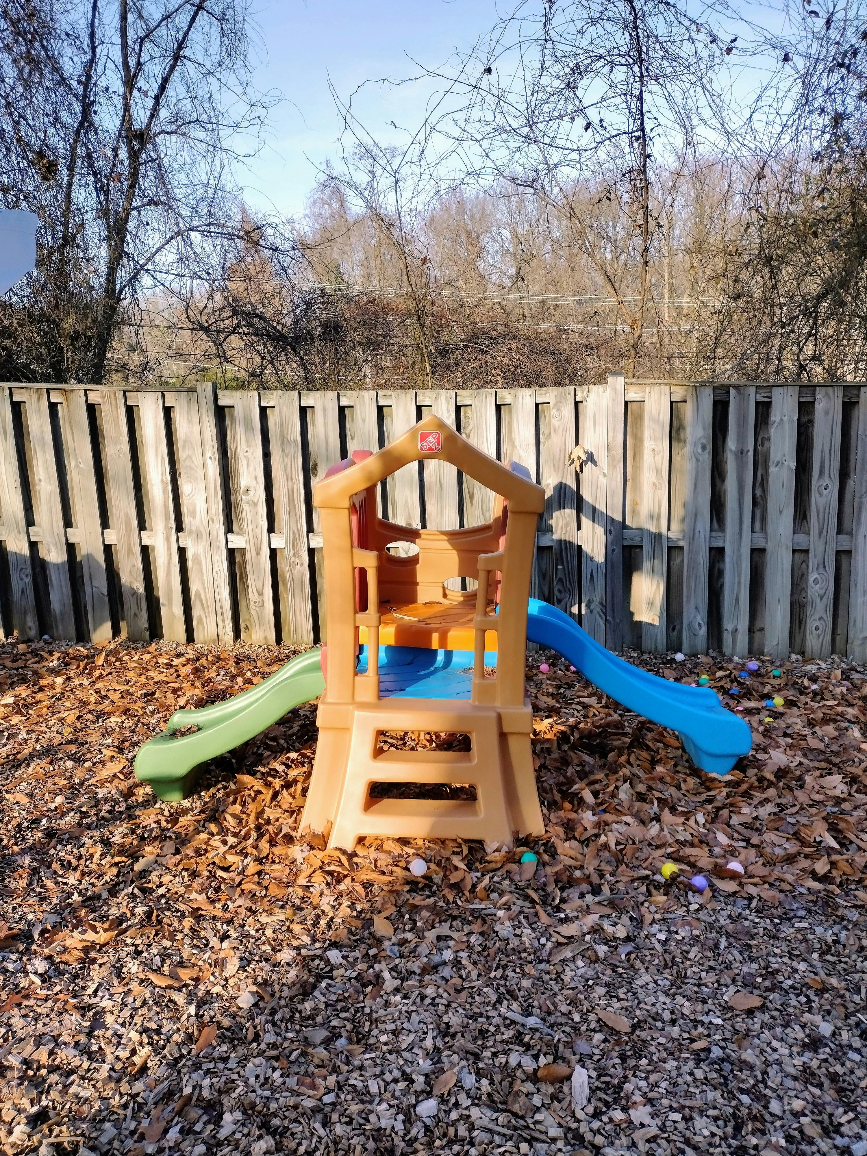 Play structure with two slides on wood chips in front of a wooden fence