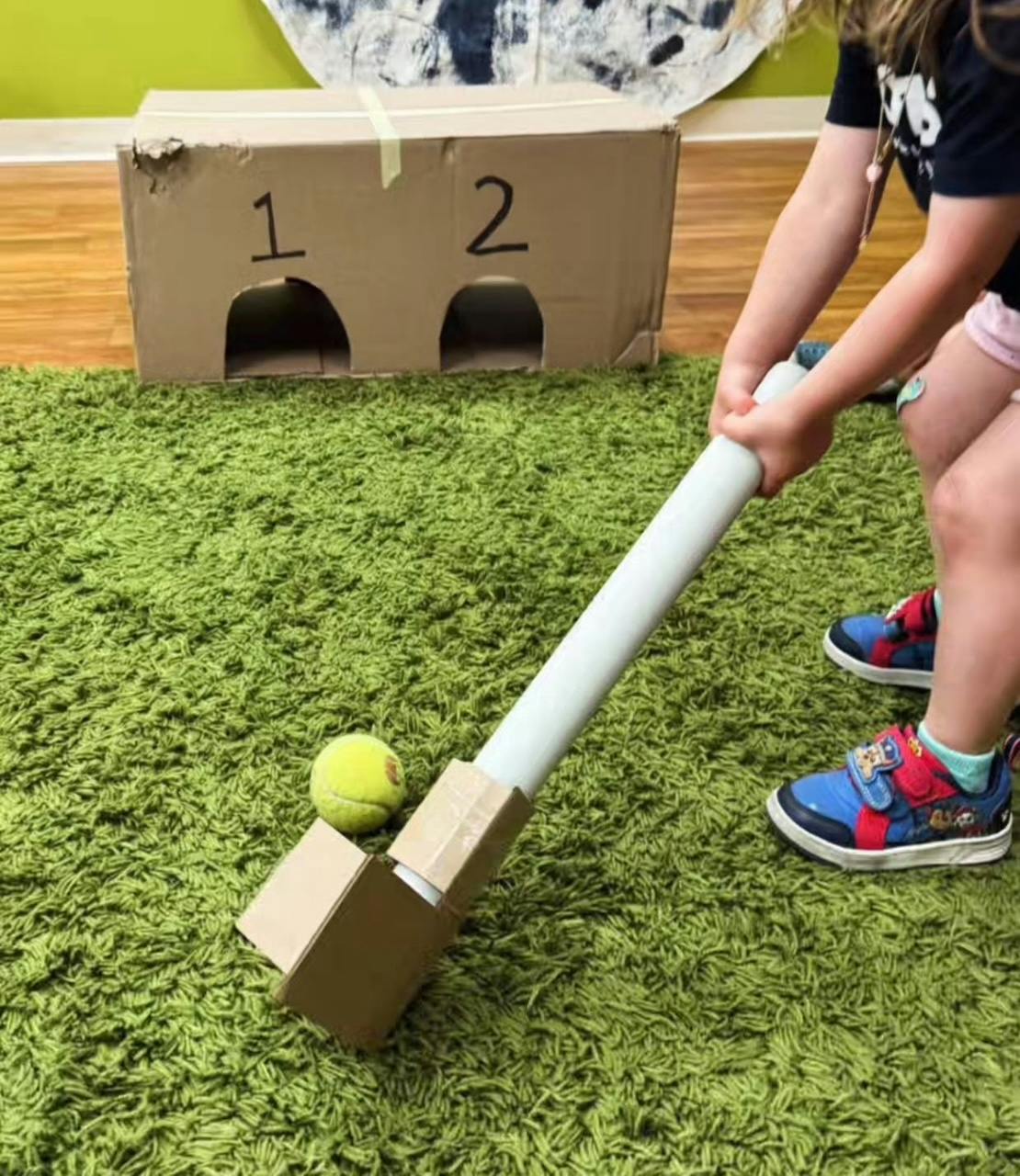 Child holding a homemade bat next to a tennis ball and cardboard box