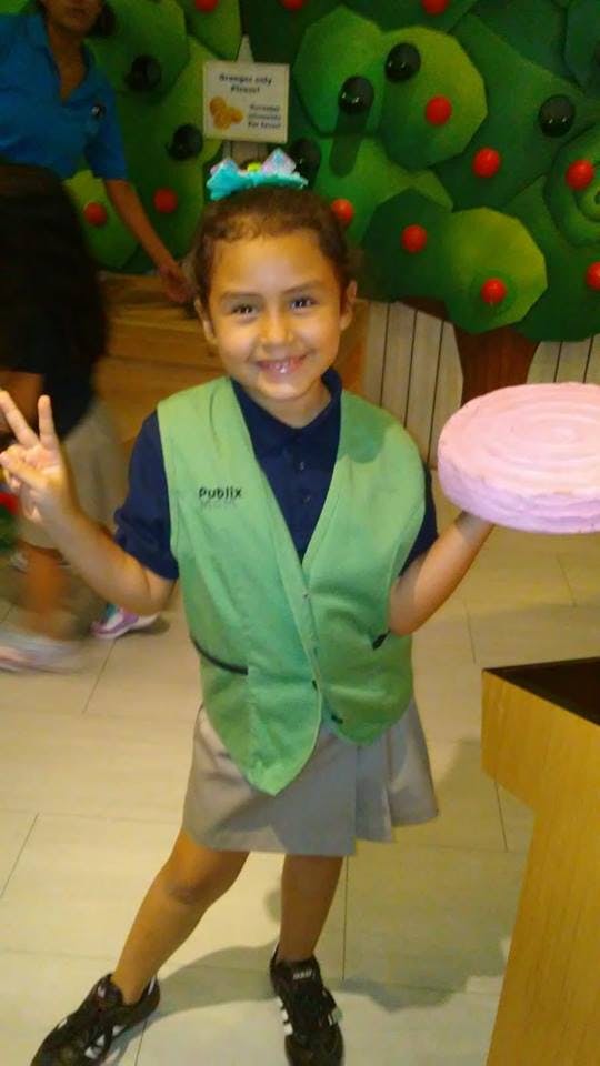 Child in green vest holding a pink object and making a peace sign