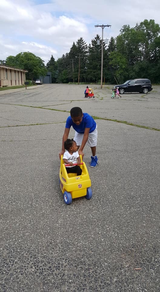 Teen pushing a child in a yellow toy car on pavement