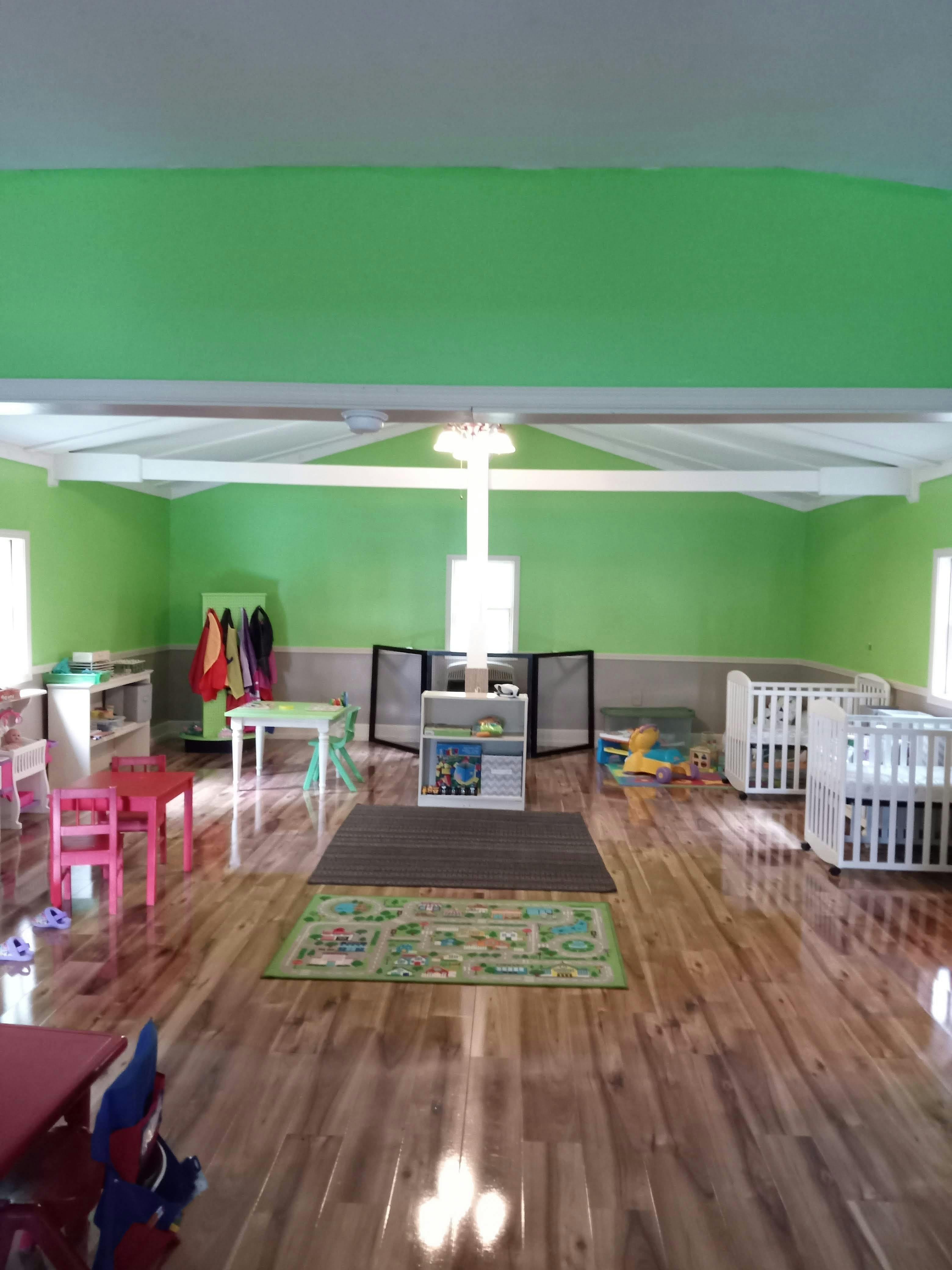 Daycare room with green walls, small tables, cribs, and play mats