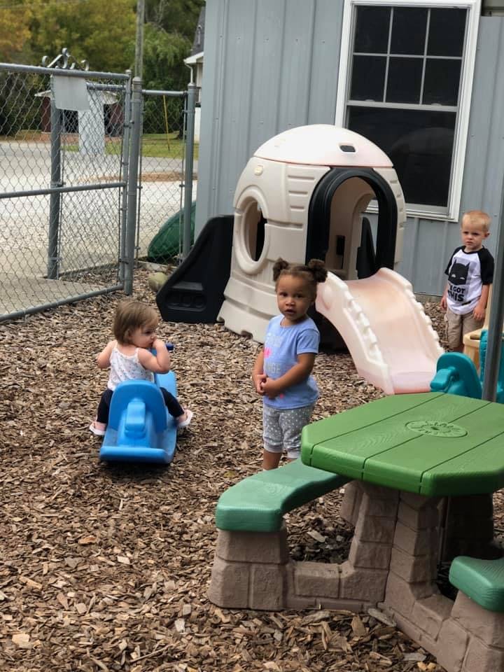 Three children on a playground with a playhouse and picnic table