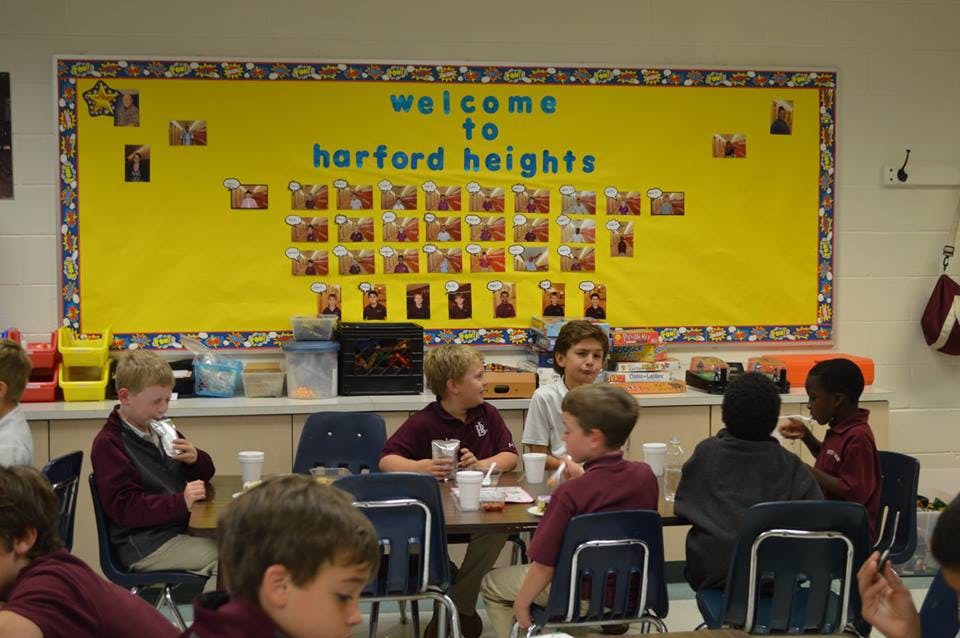 Children sitting at tables in a classroom with a "Welcome to Harford Heights" sign