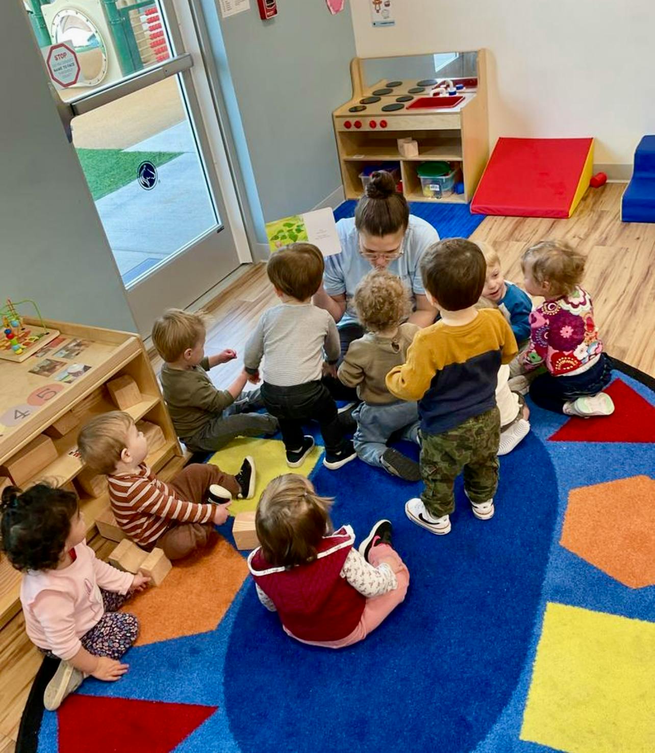 Adult reading a book to a group of children sitting on a colorful playroom rug