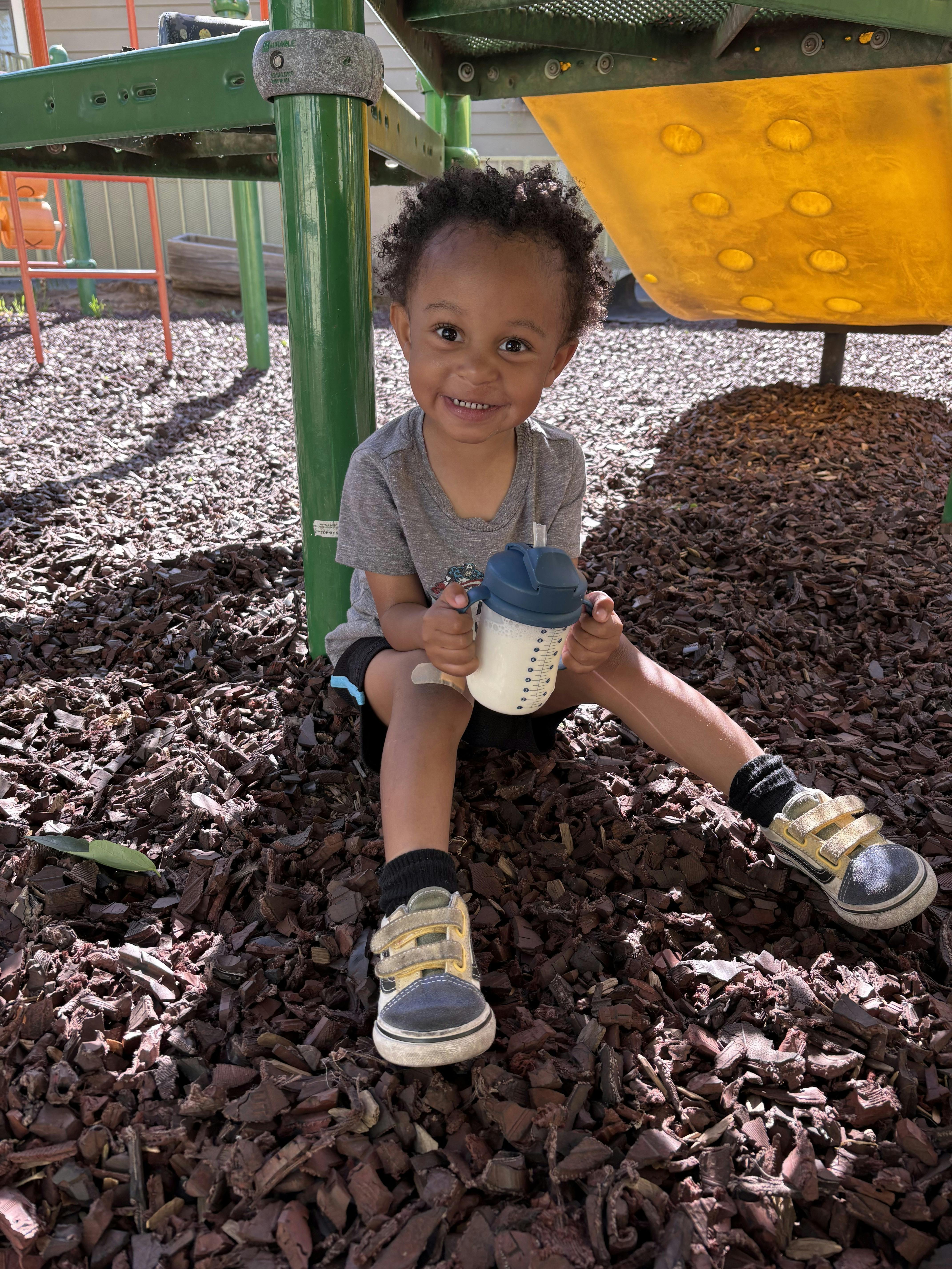 Child sitting on wood chips at a playground, holding a sippy cup