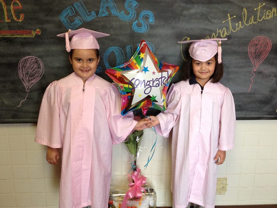 Two children in pink graduation gowns holding a star-shaped balloon that says "Congrats"