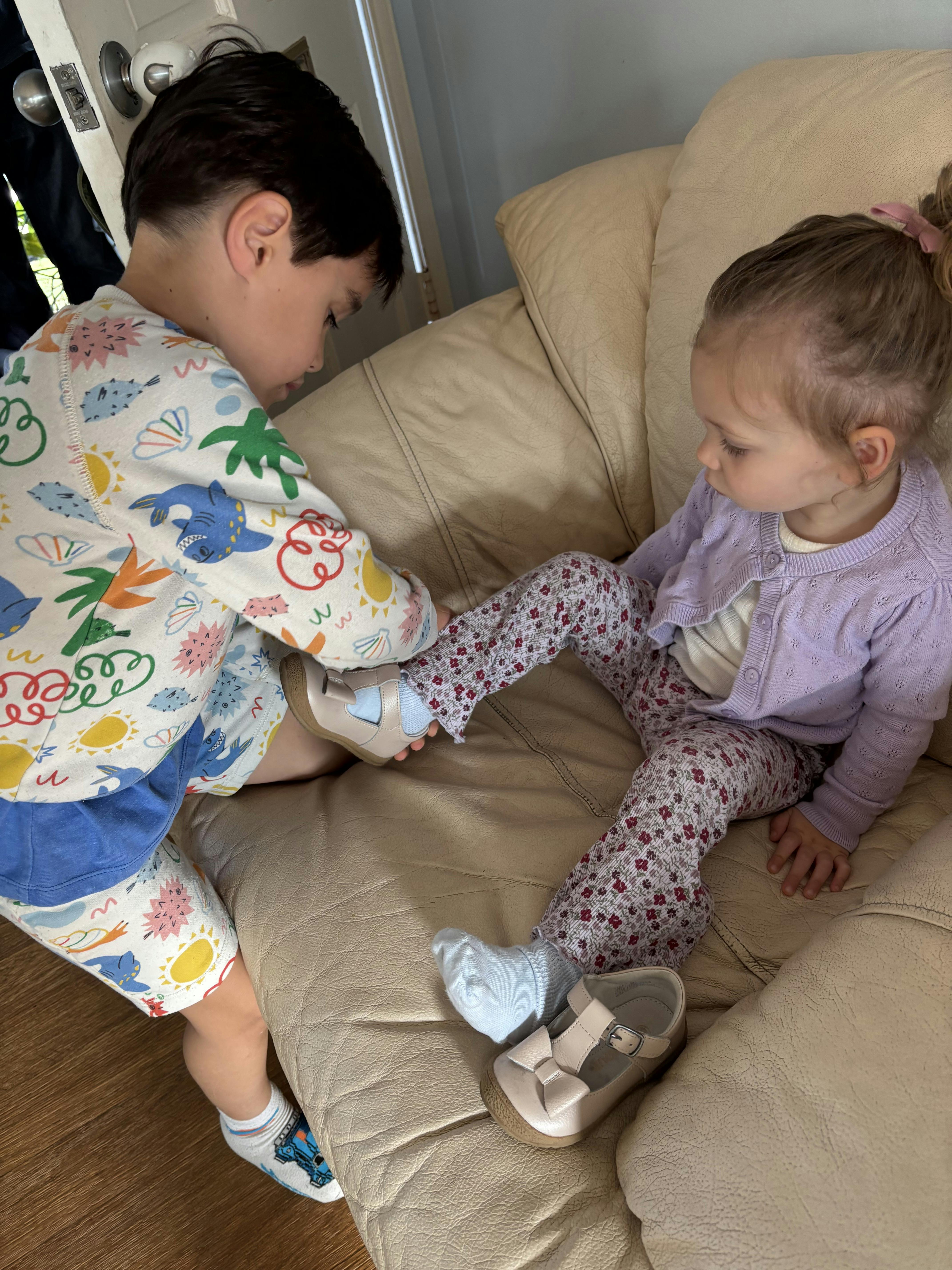 Child helping another child put on a shoe on a light brown couch