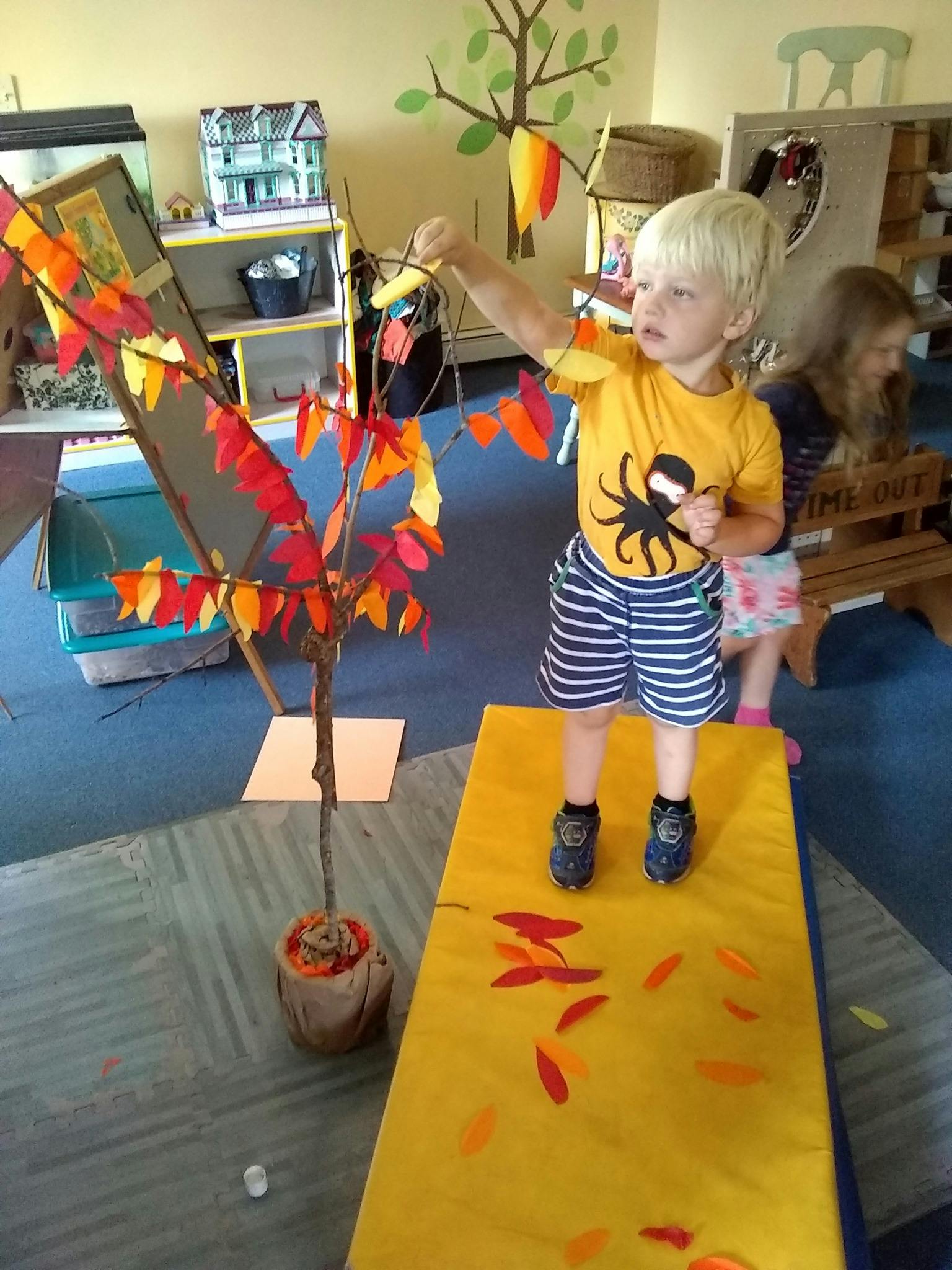 Child standing on a yellow mat, reaching for a decorative tree with colorful leaves