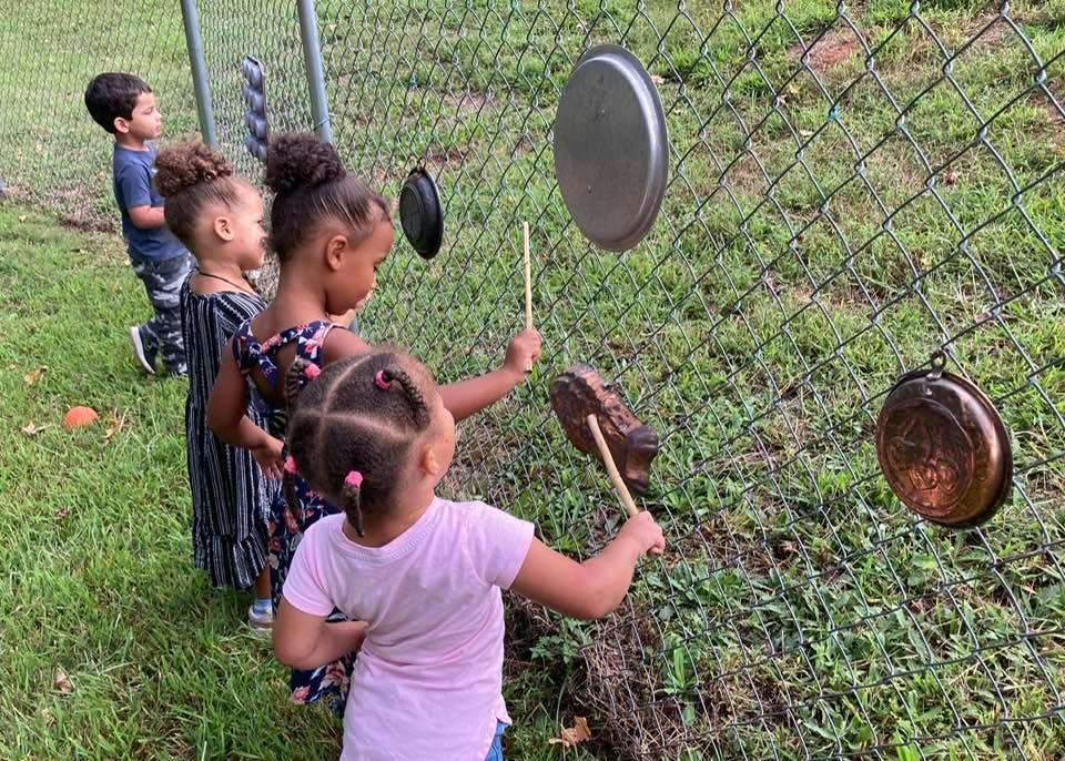 Children playing music on pots and pans hanging from a chain link fence