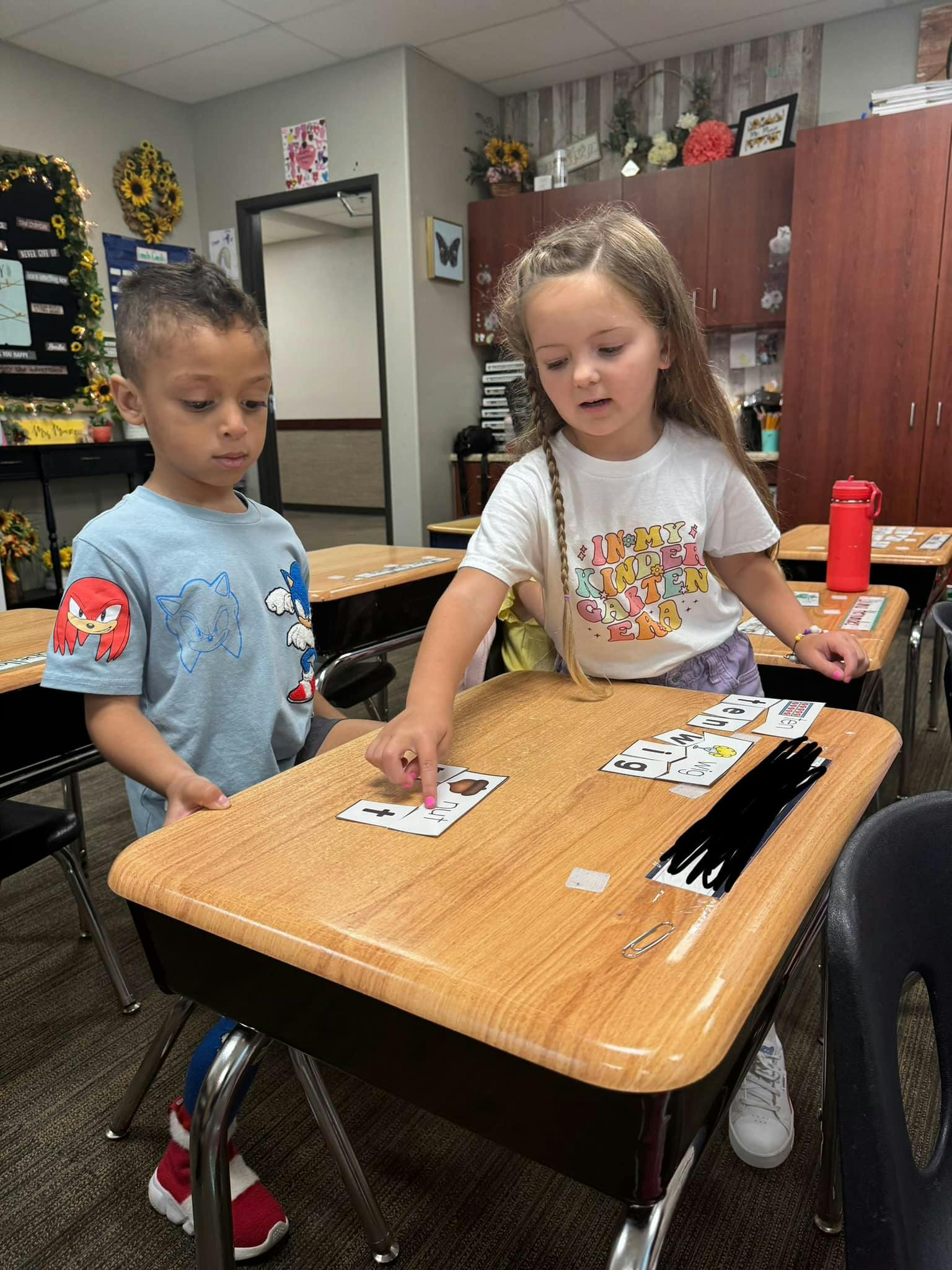 Two children standing at a desk, one pointing at a card