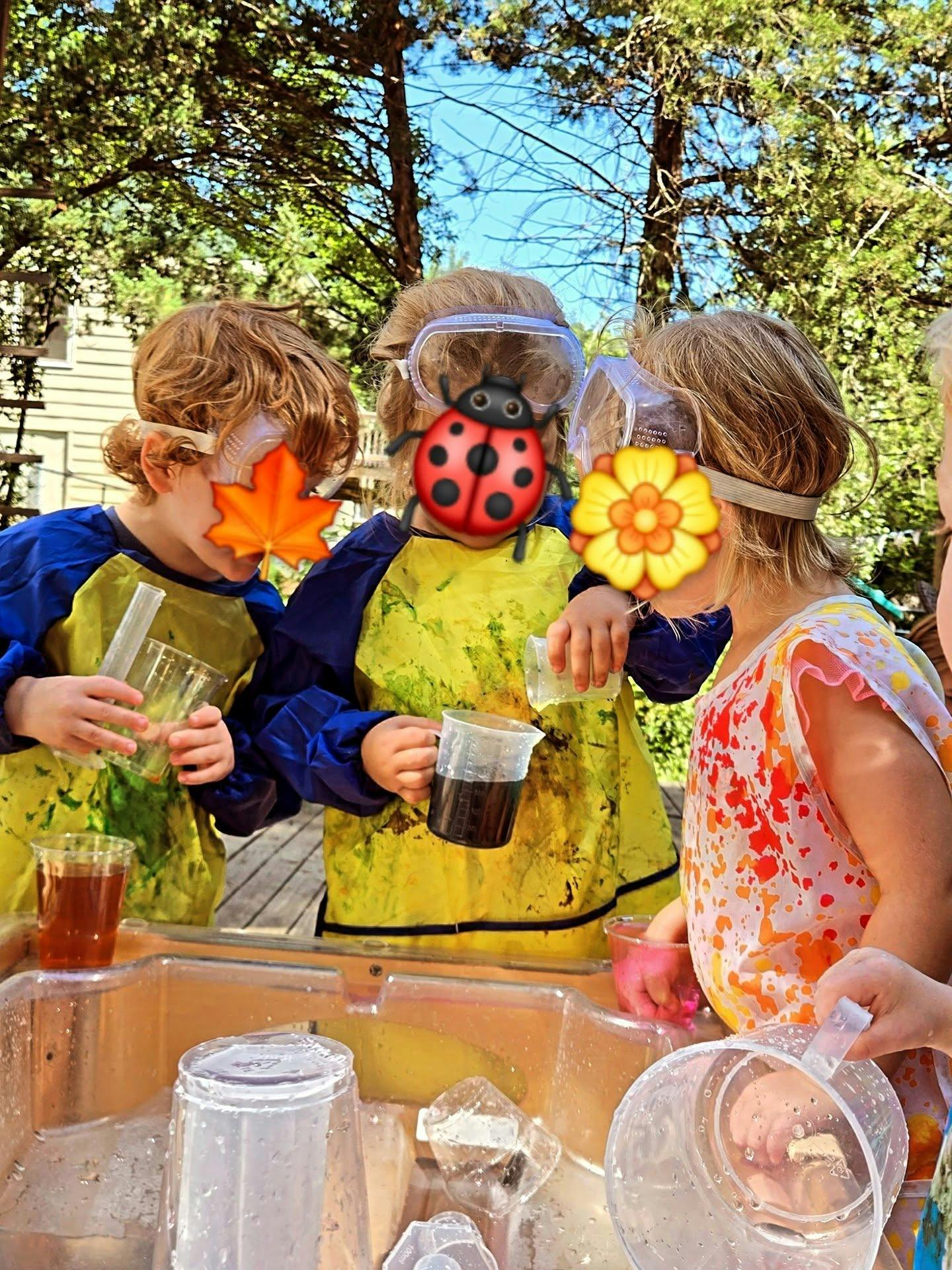 Three children wearing goggles and smocks playing with water and plastic beakers