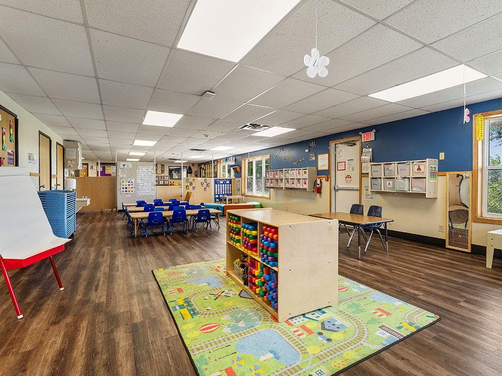 Empty classroom with small tables and chairs, a toy shelf, and a playmat