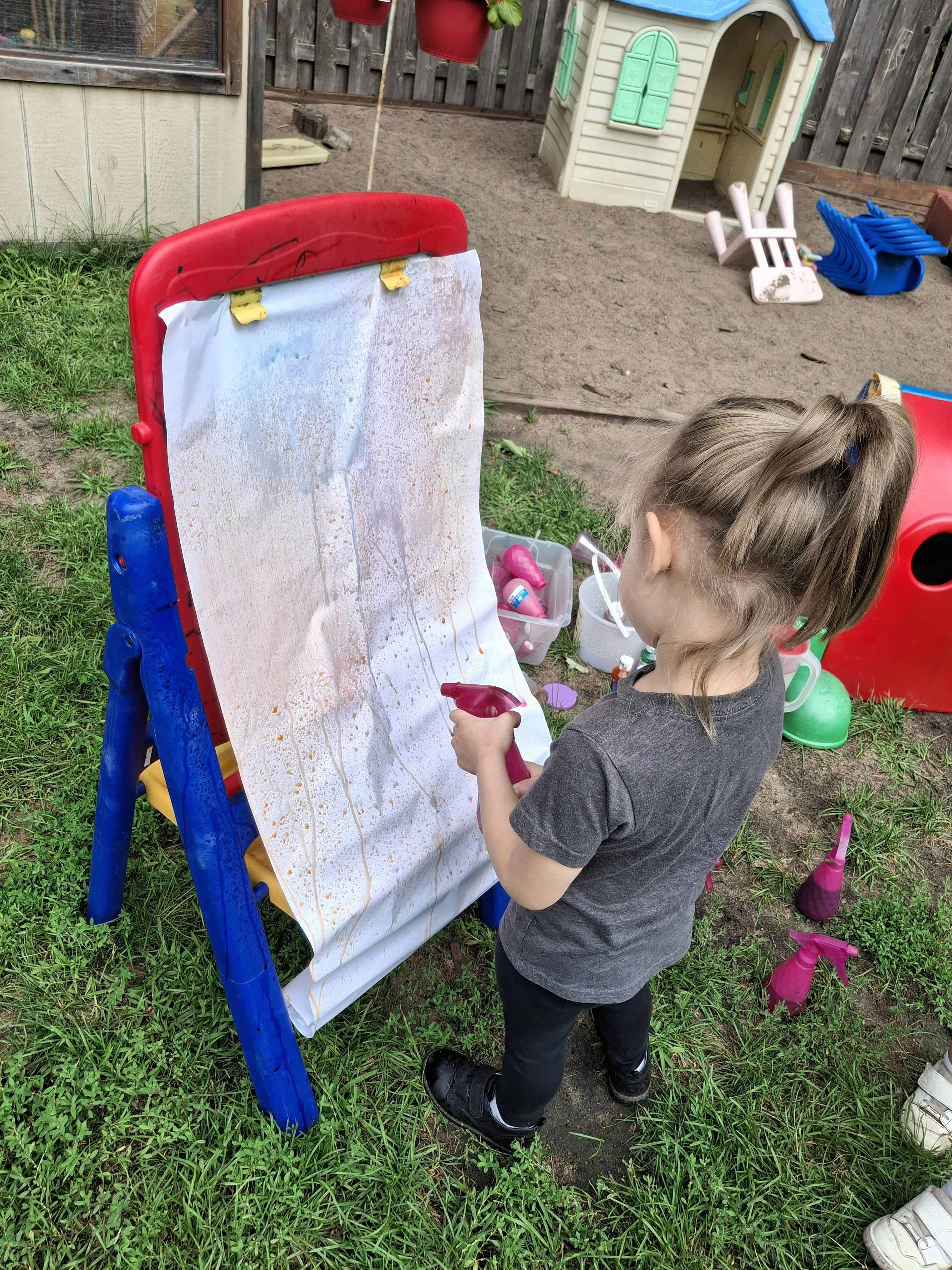 Child spraying water onto a white sheet on an easel outdoors