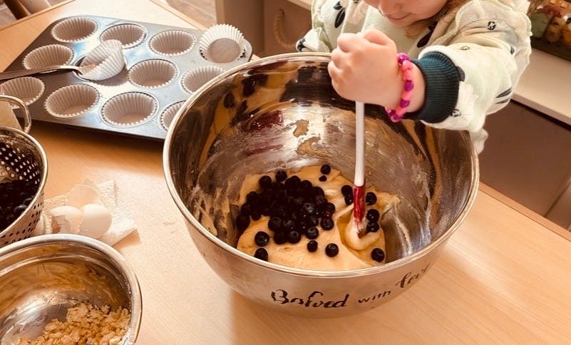 Child mixing blueberries into batter in a large bowl, muffin tin in background
