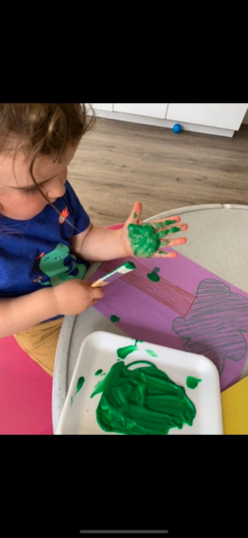 Child with green paint on hand holding a paintbrush over paper