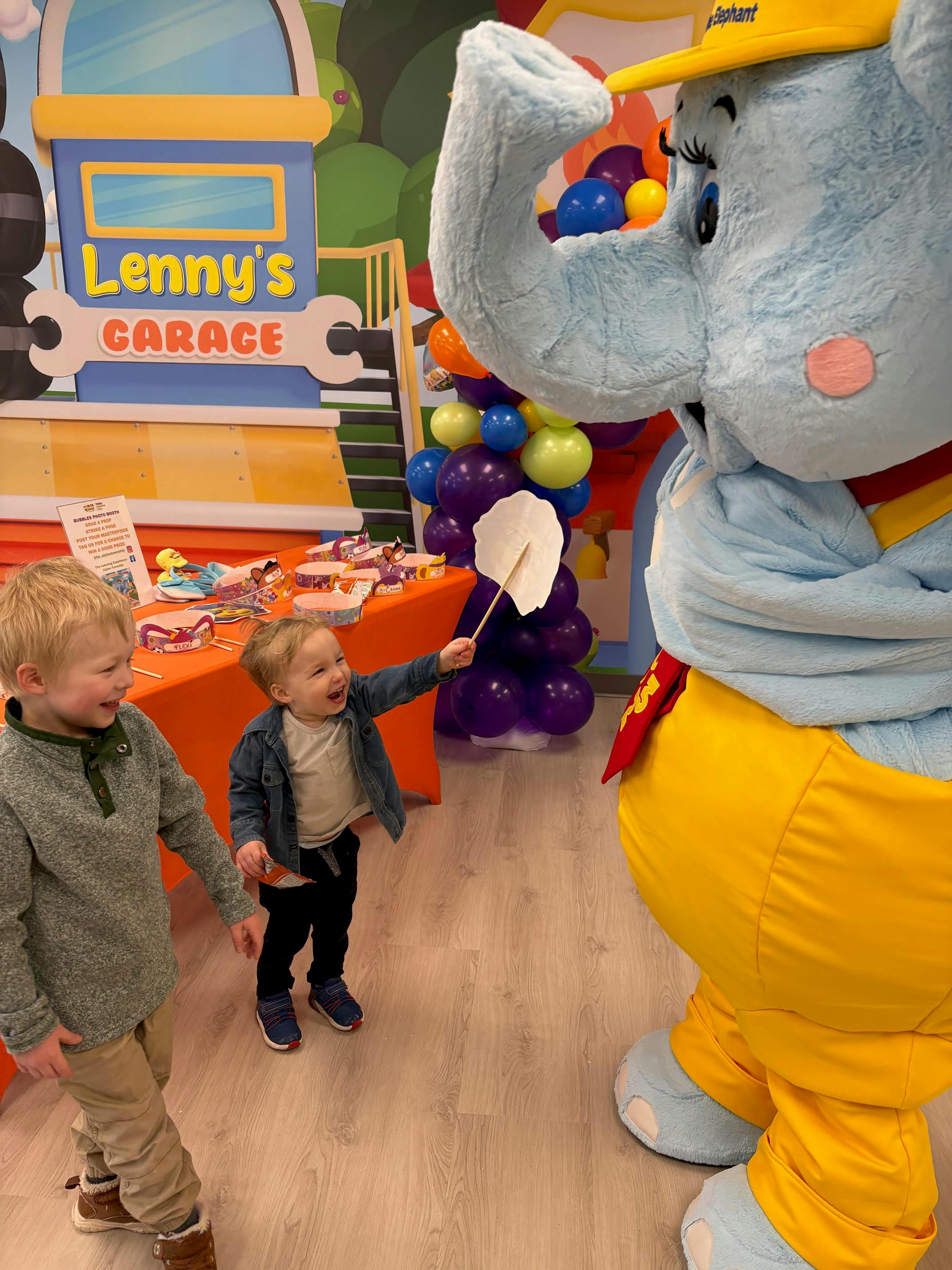 Child offering a paper fan to an elephant mascot, another child looks on