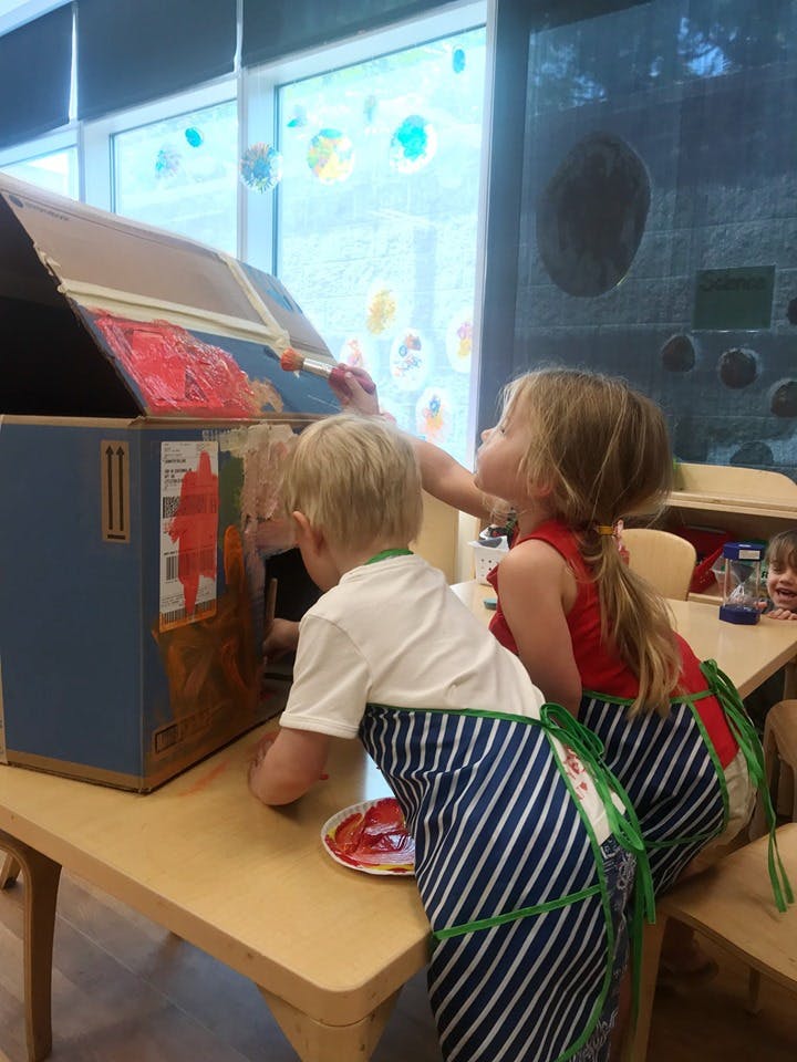 Two children painting a cardboard box in a classroom
