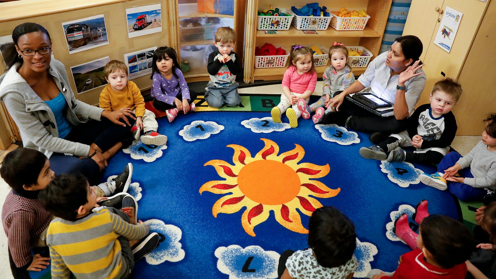 Two adults and multiple children sitting on a sun-patterned rug in a classroom