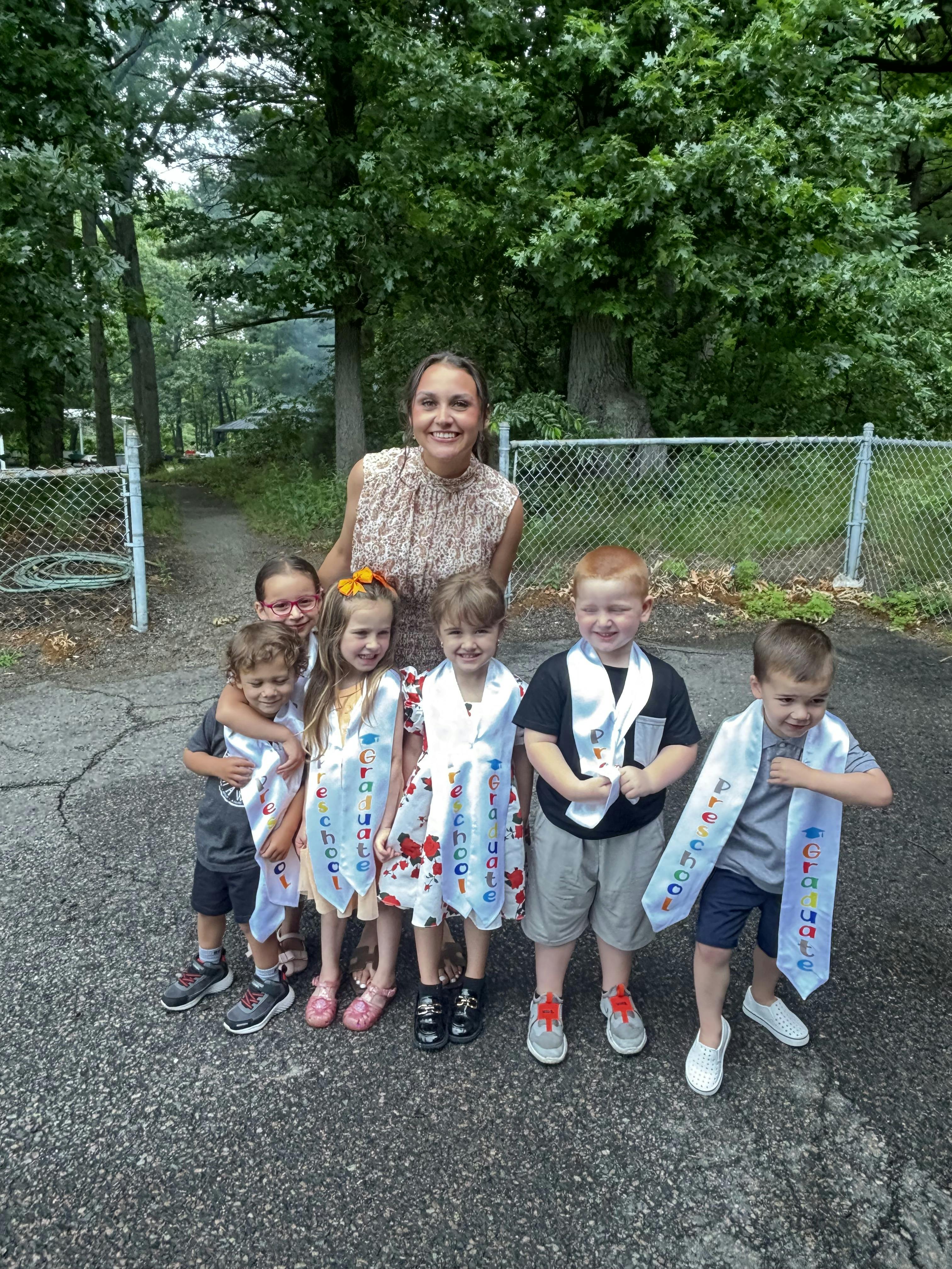 Adult and several children wearing preschool graduate sashes standing outdoors