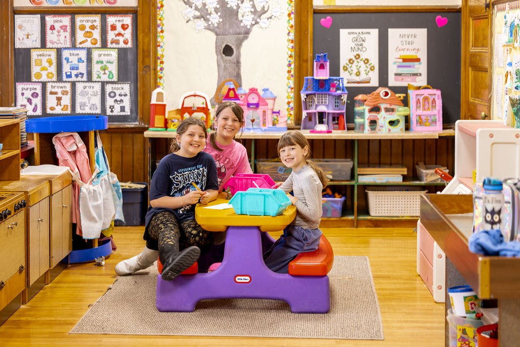 Three children smiling at a small table in a classroom