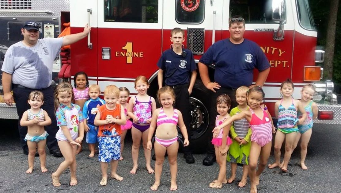Three people and multiple children in swimsuits in front of a fire truck