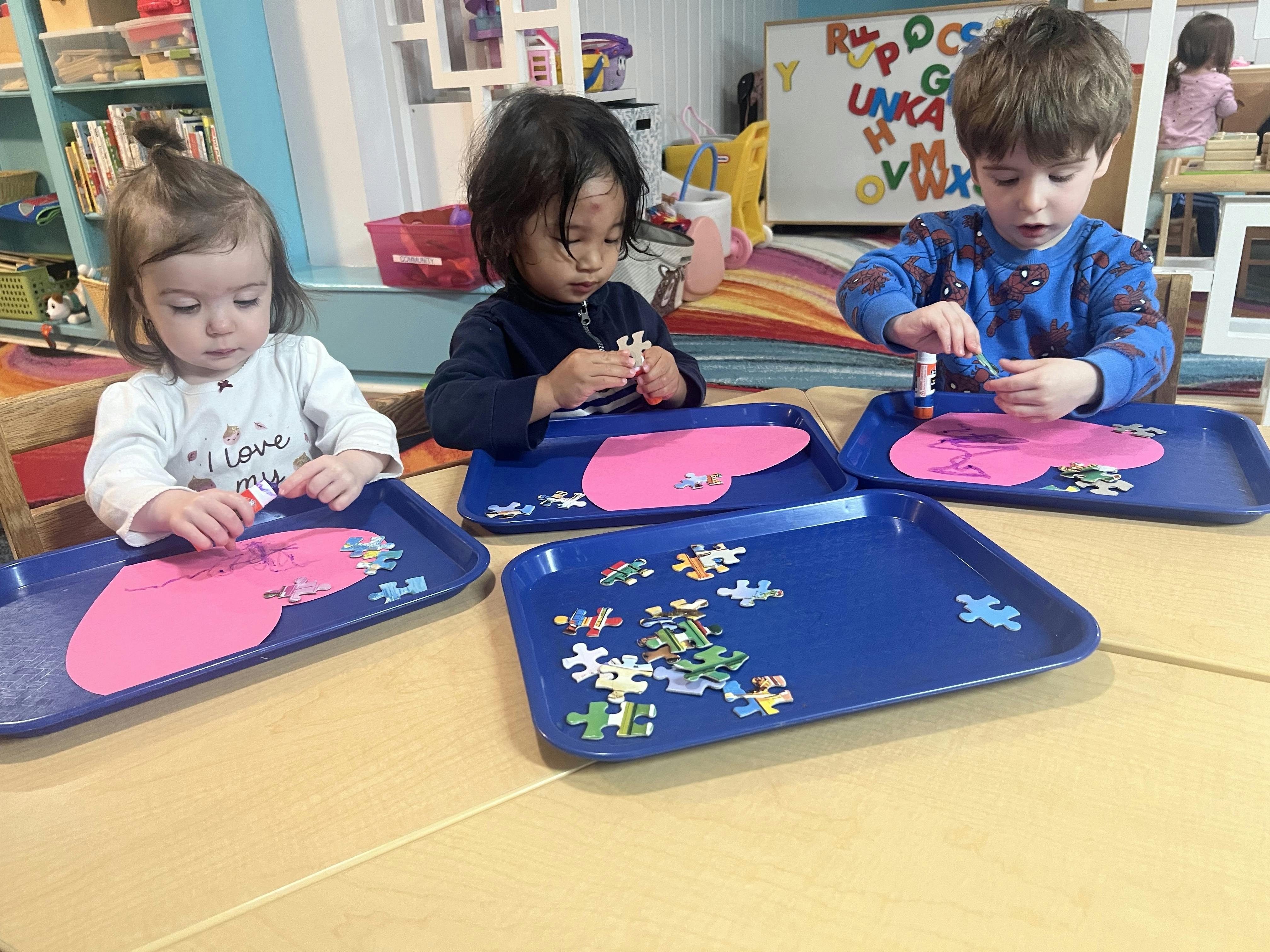 Three children working on heart-themed craft projects at a classroom table
