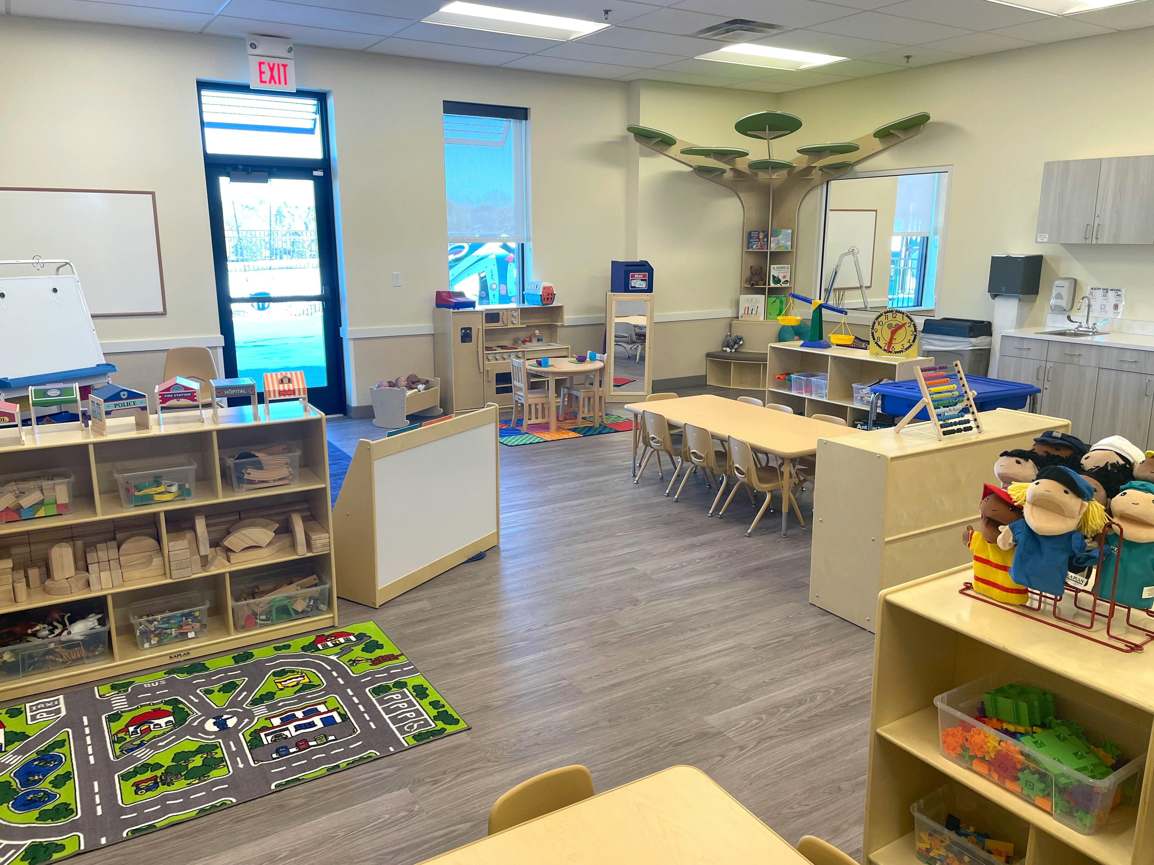 A classroom with small tables and chairs, shelves with toys, and a play rug