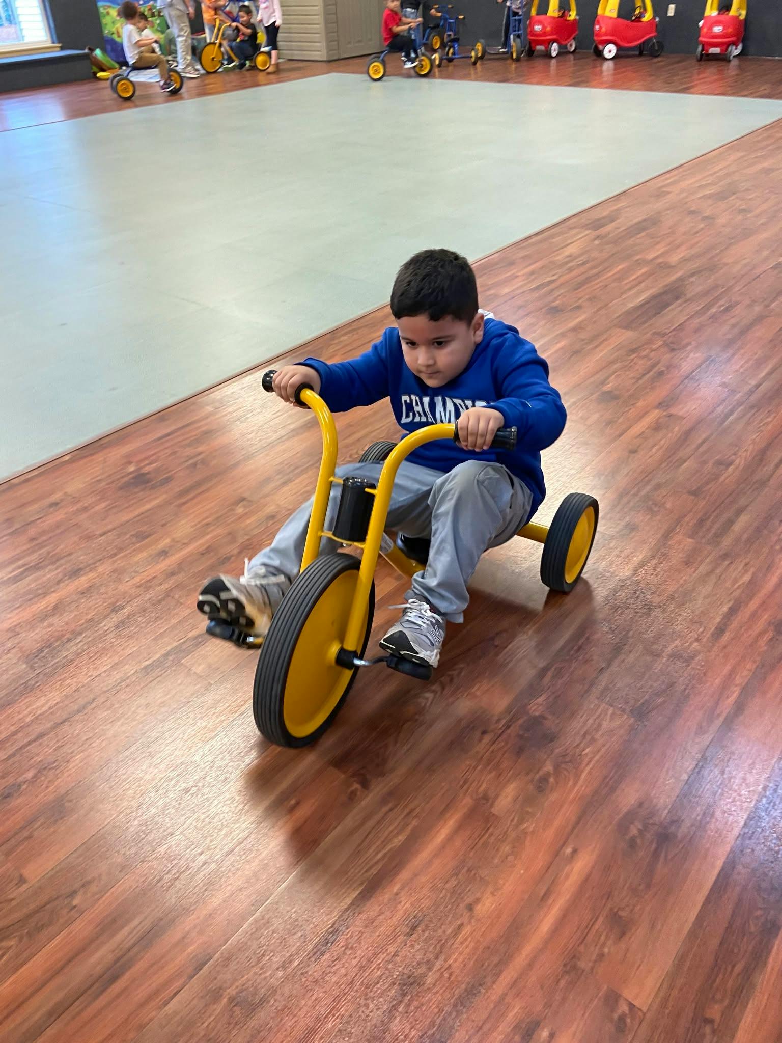 Child riding a yellow tricycle on a wood floor in a playroom