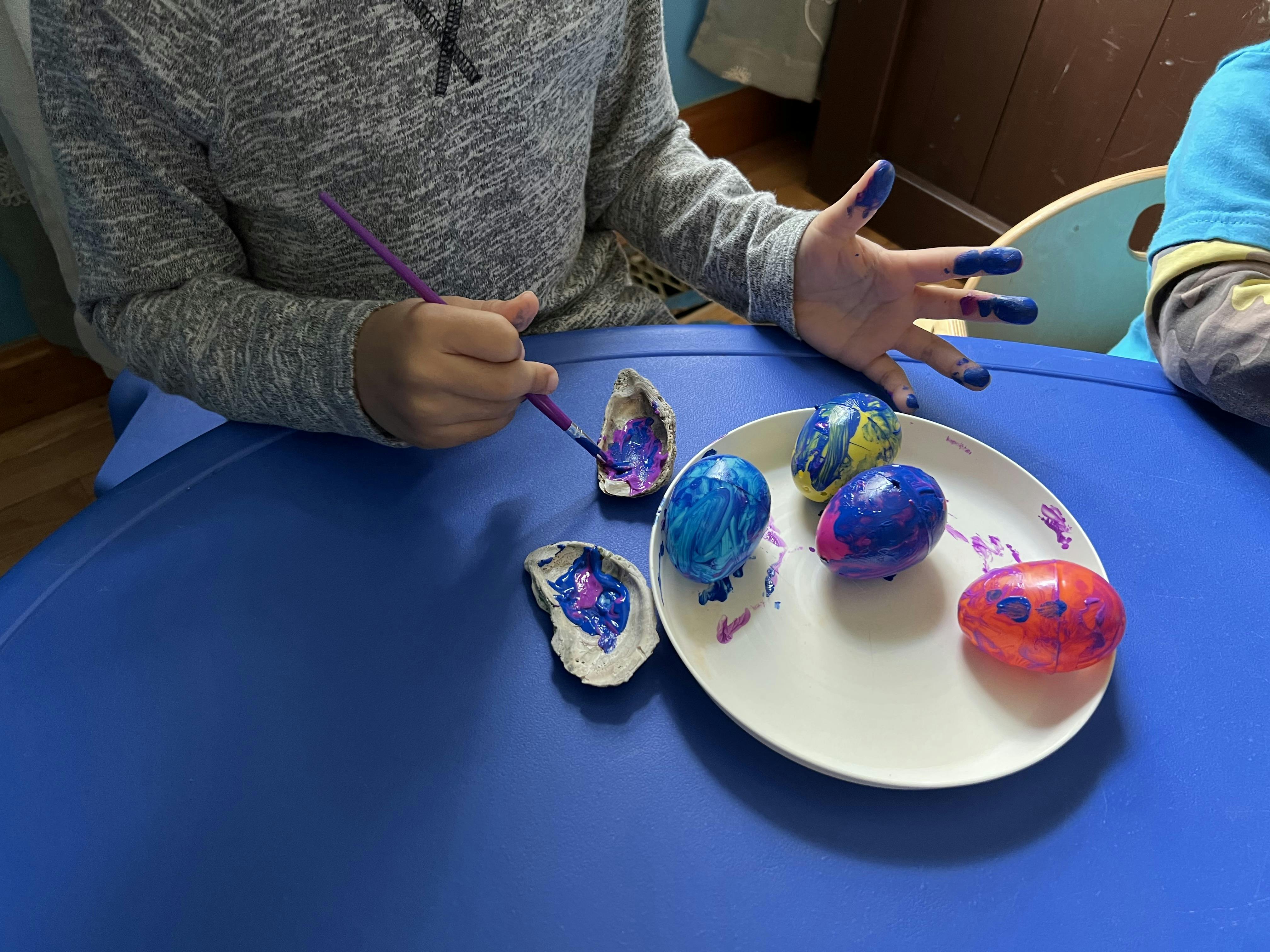 Child painting shells and eggs with a brush on a blue table