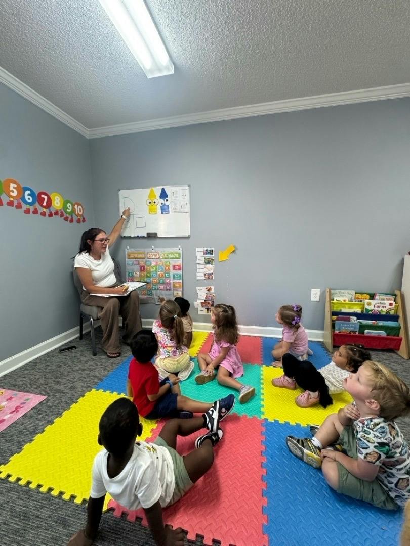 Teacher pointing to a whiteboard while children sit on a colorful floor mat