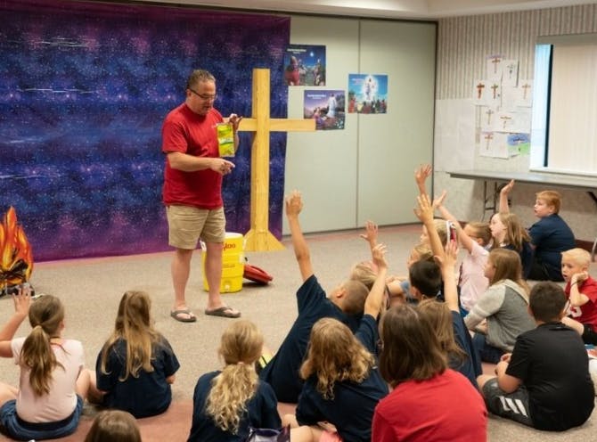 Adult speaking to children sitting on floor, many with hands raised