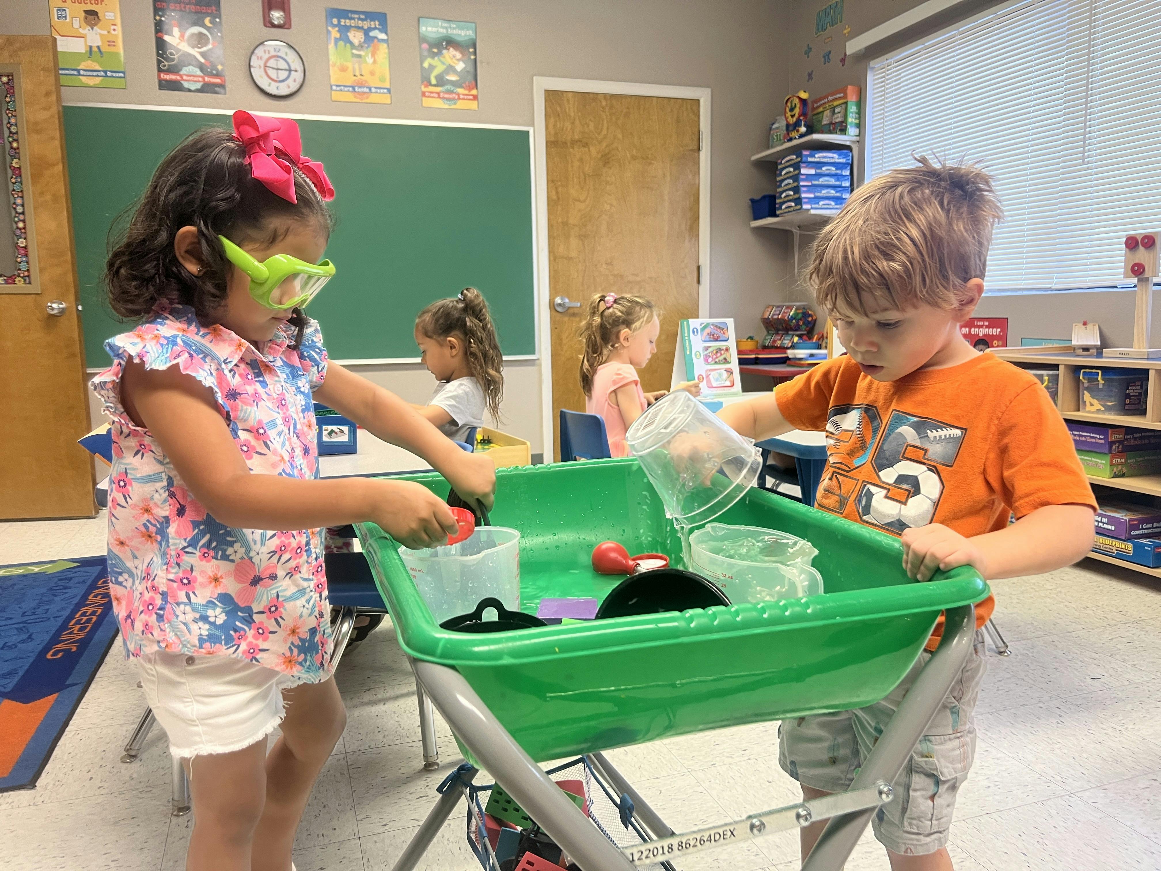 Two children playing with water and containers at a green table