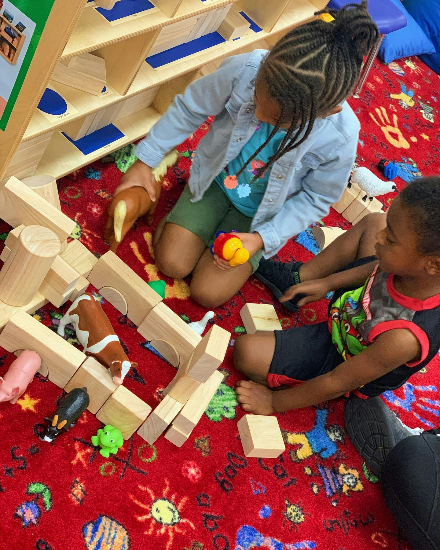 Two children playing with wooden blocks and animal toys on a red rug