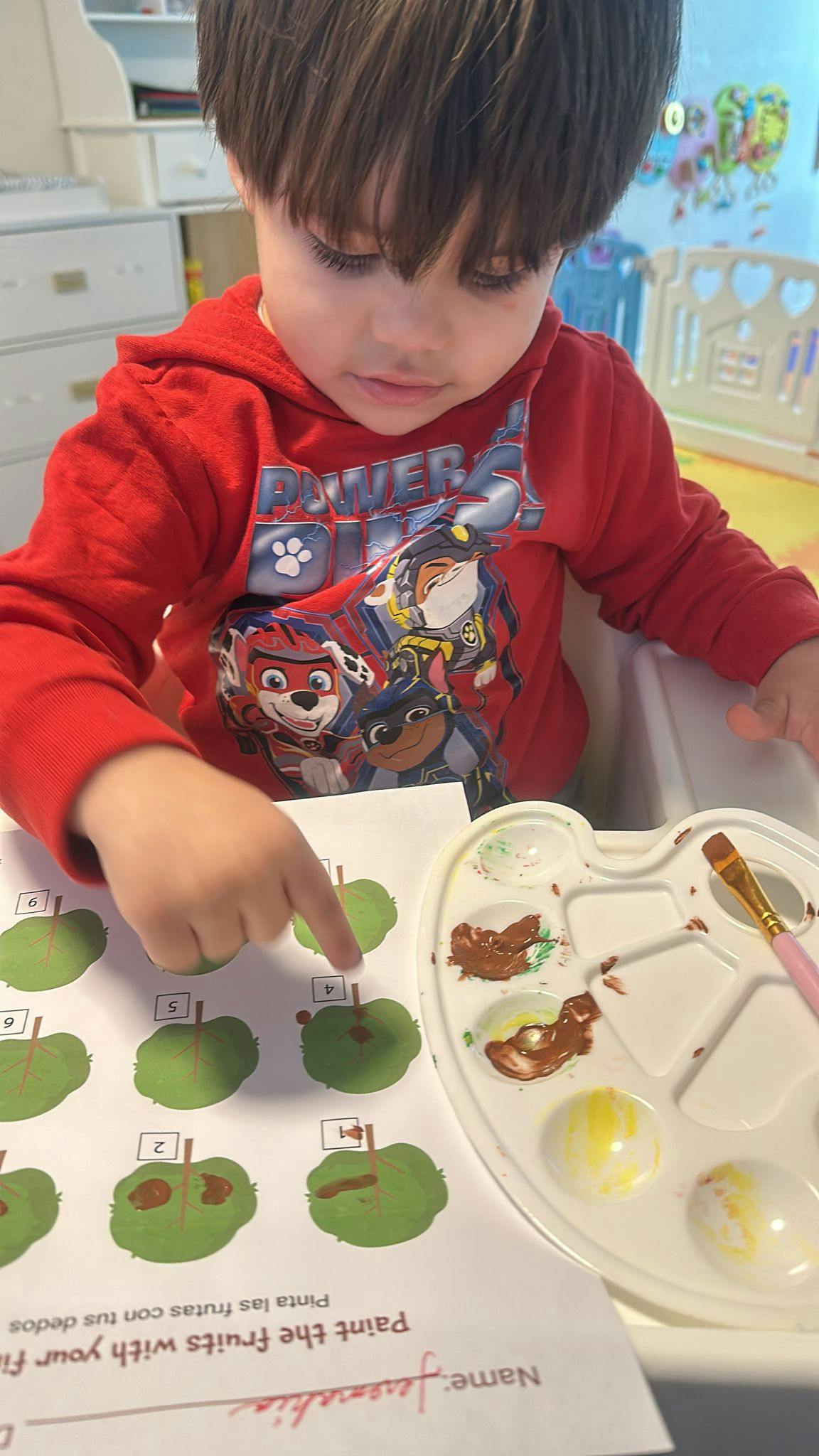 Child pointing at a paper with green apple shapes and a paint palette