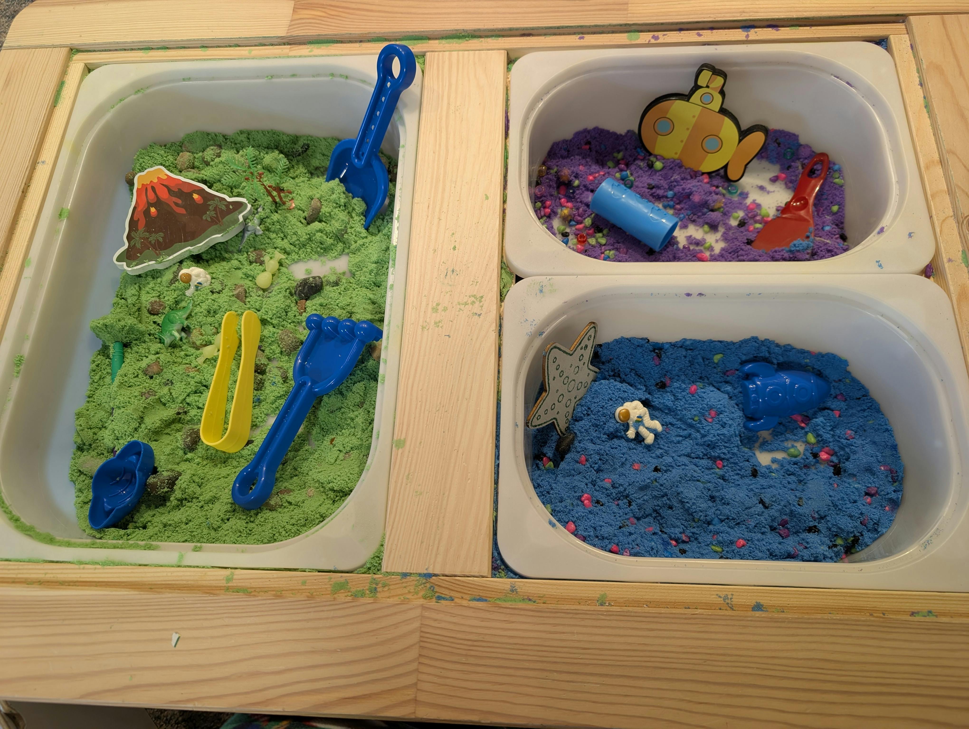 Sensory bin with green sand and volcano, purple sand with submarine, blue sand with astronaut