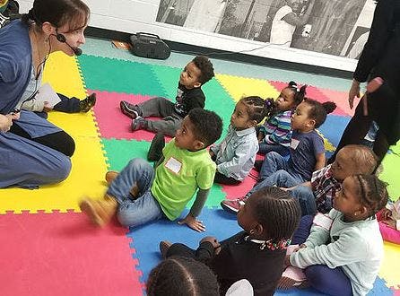Person with headset speaking to a group of children sitting on a colorful mat
