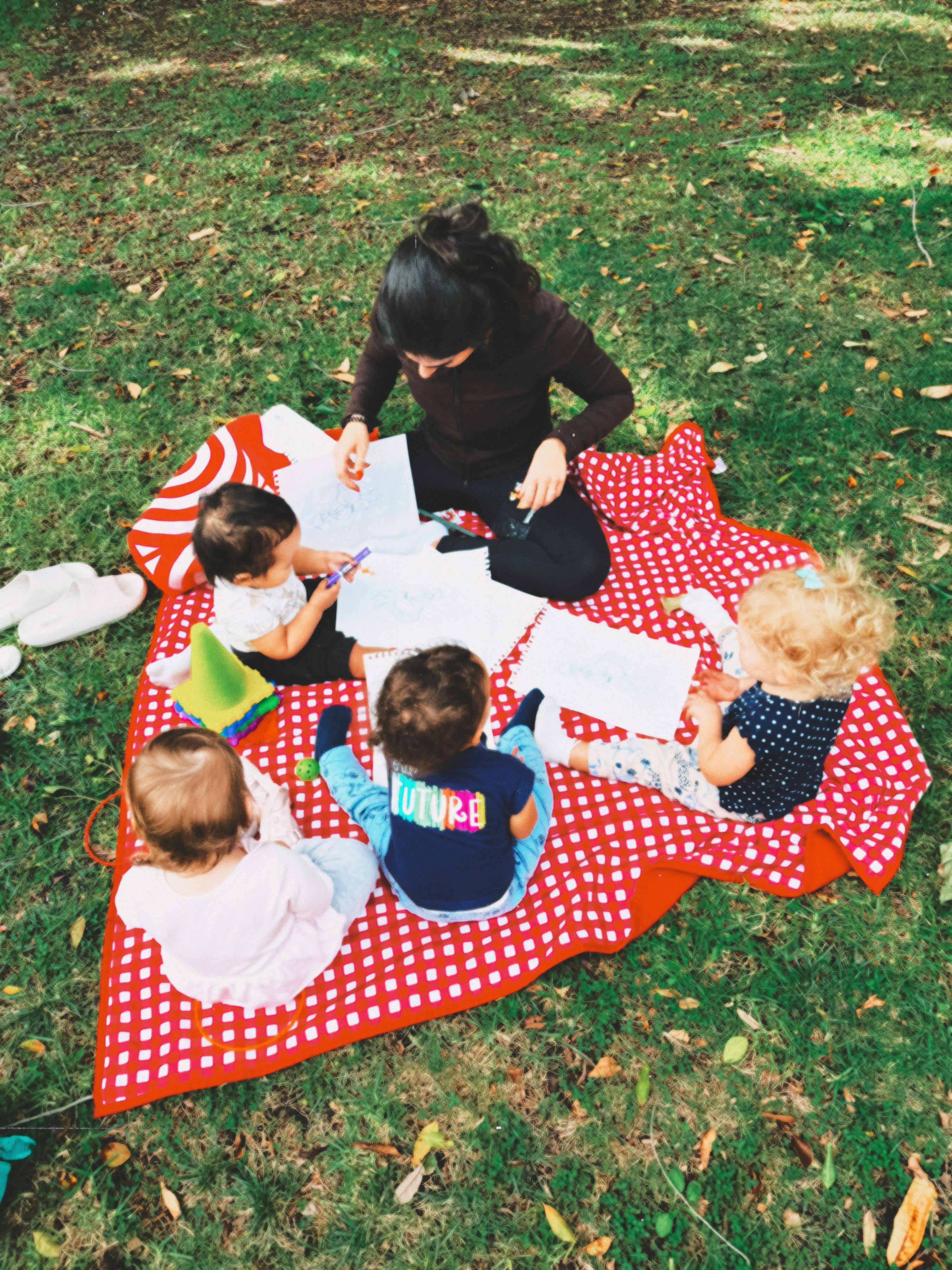 Woman and four children drawing on a red and white blanket on grass
