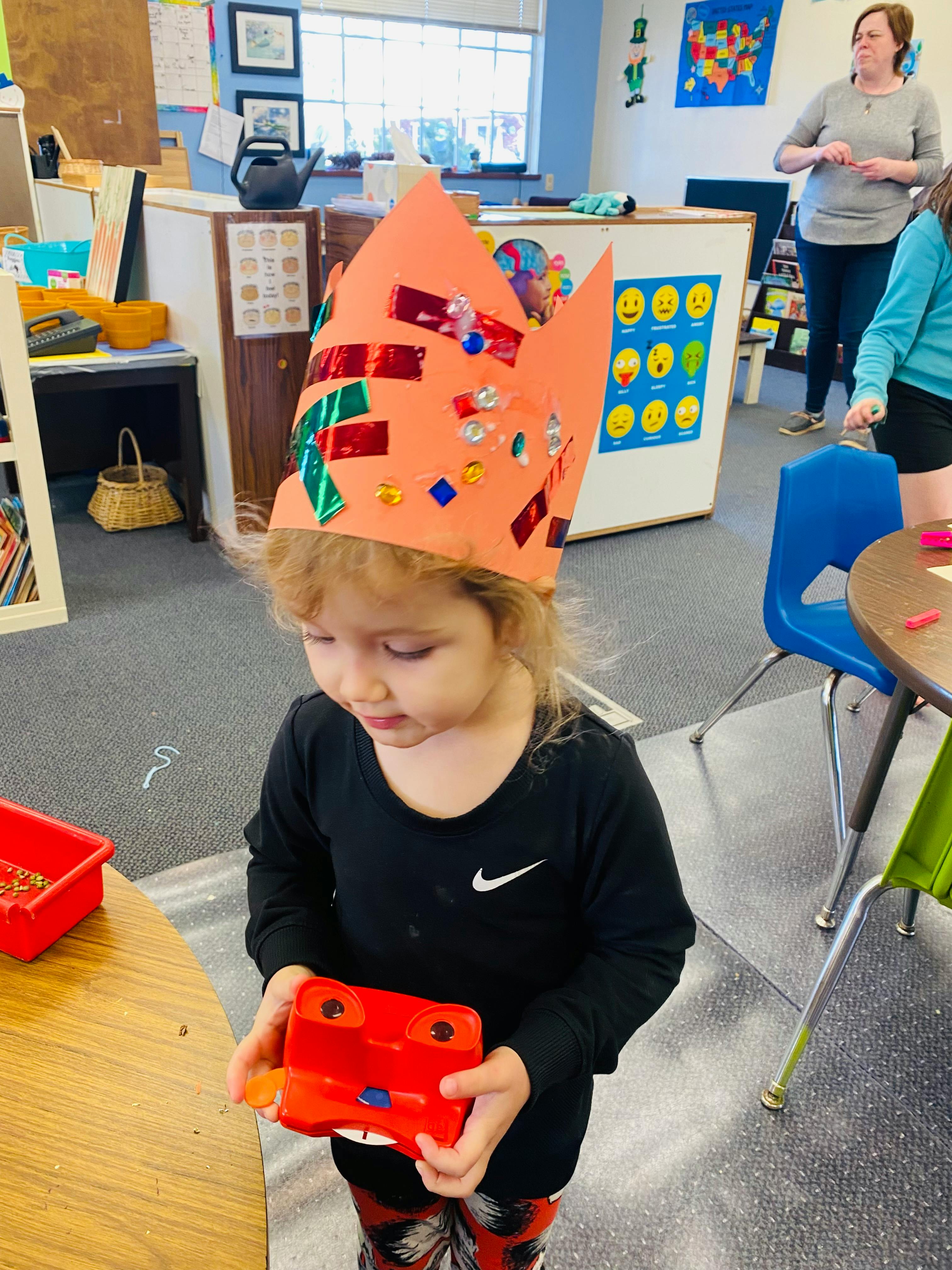 Child wearing a decorated paper crown and holding a red toy