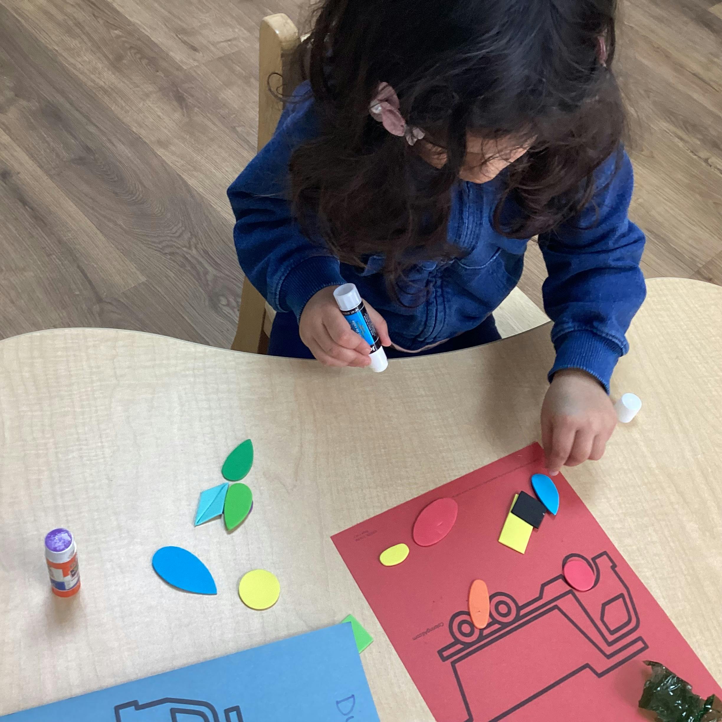 Child gluing paper shapes onto a red paper with a truck outline