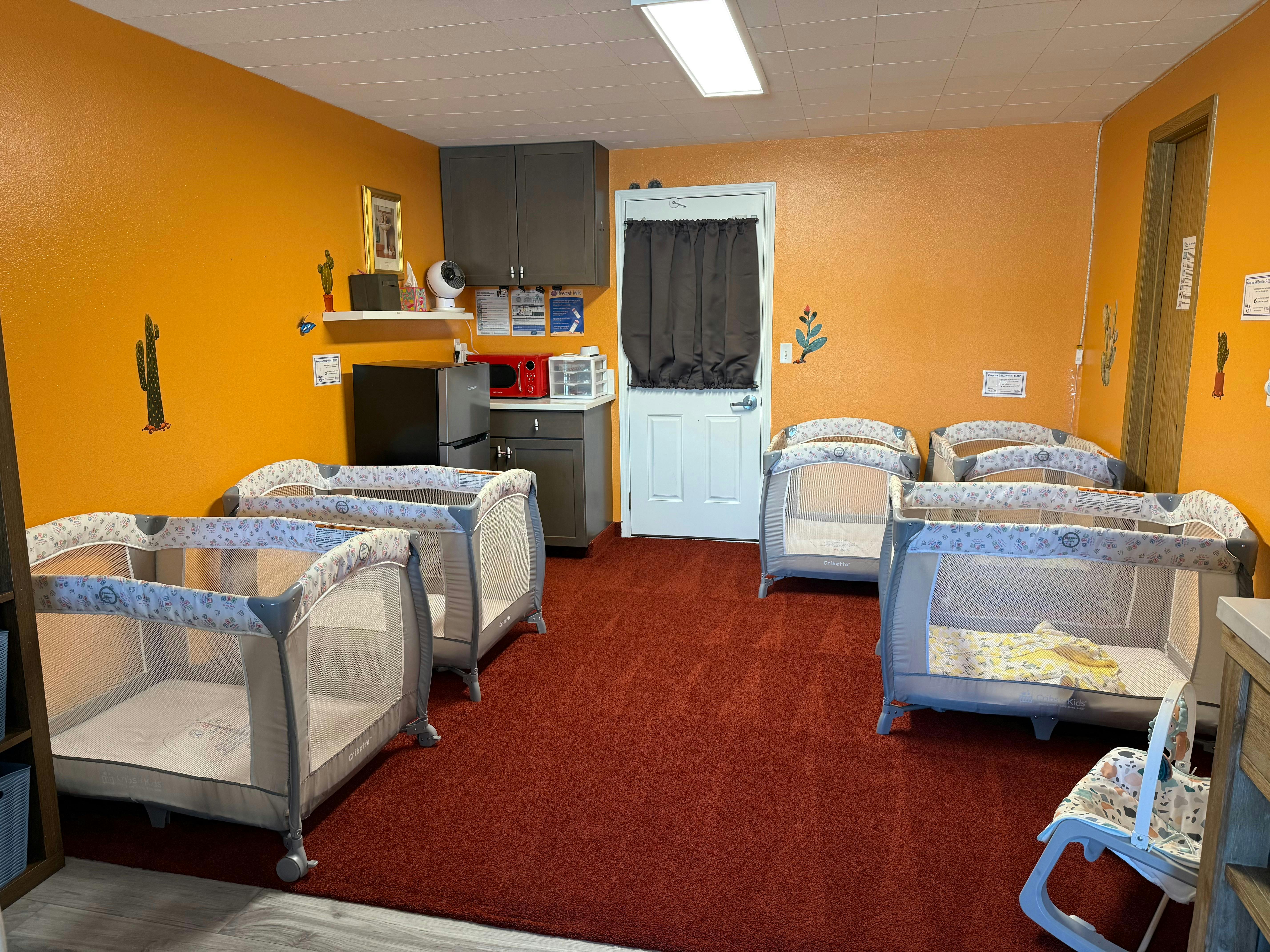 Multiple empty cribs in a room with orange walls and red carpet