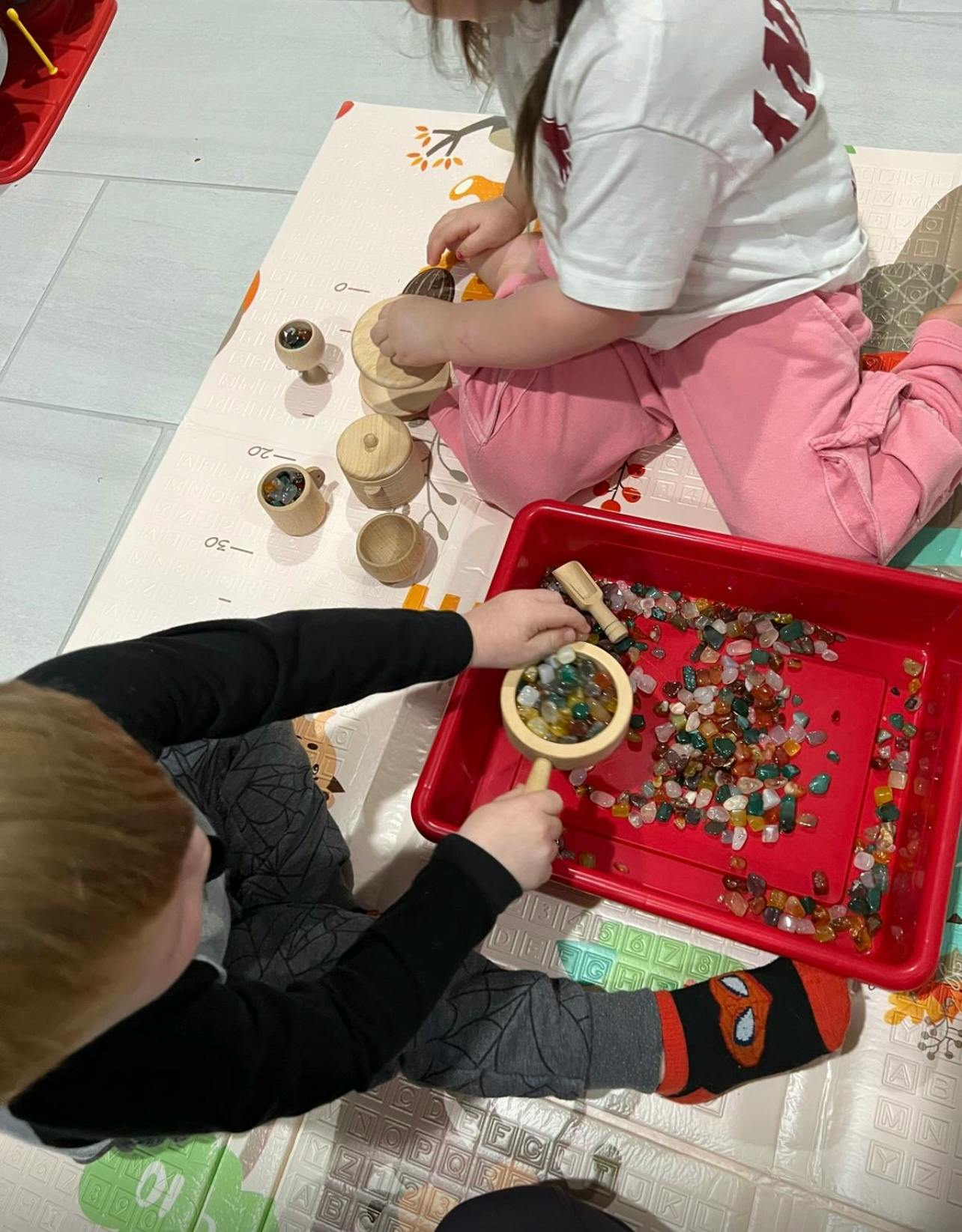 Two children playing with wooden toys and colorful stones in a red tray