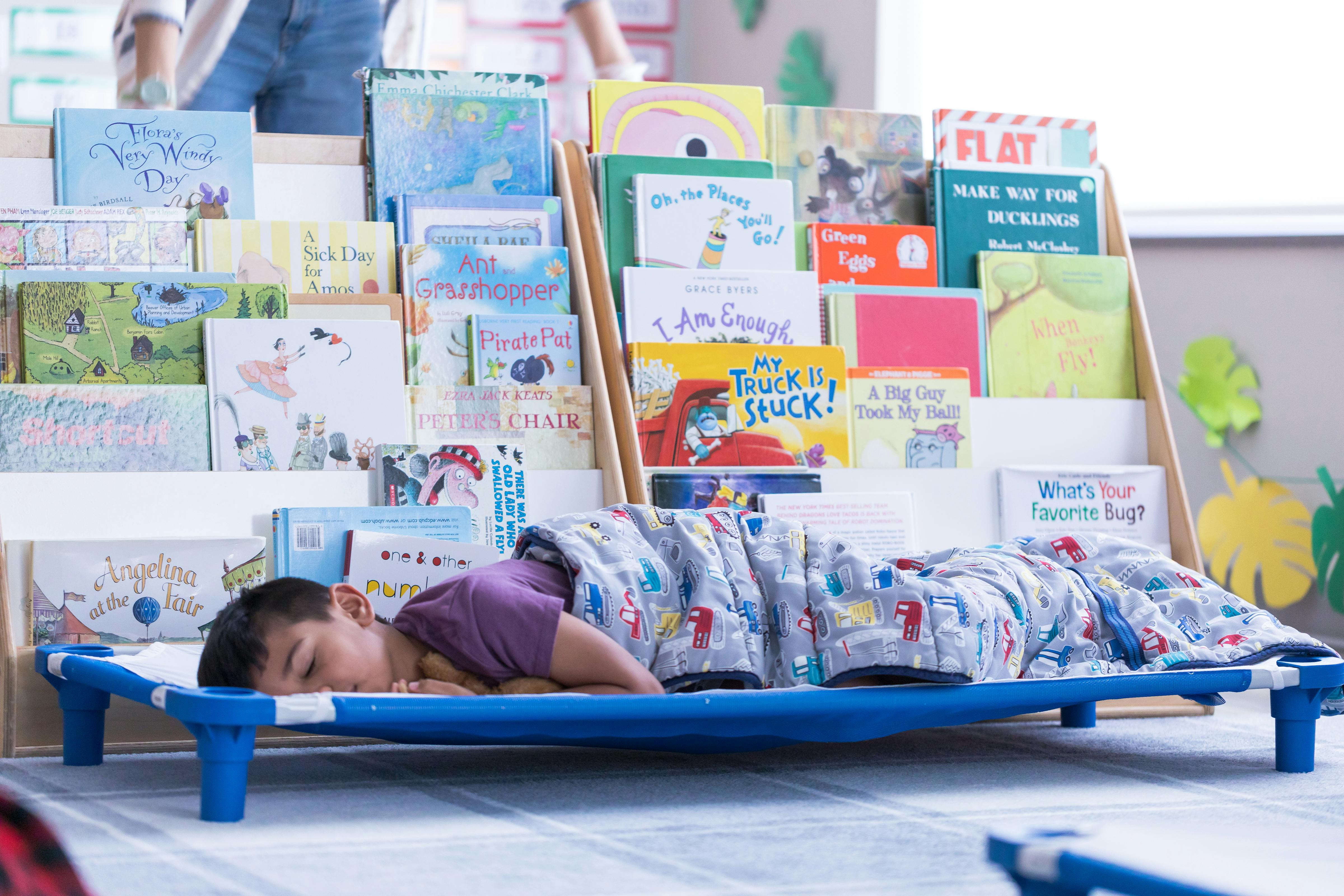 Child sleeping on a cot in front of bookshelves filled with books