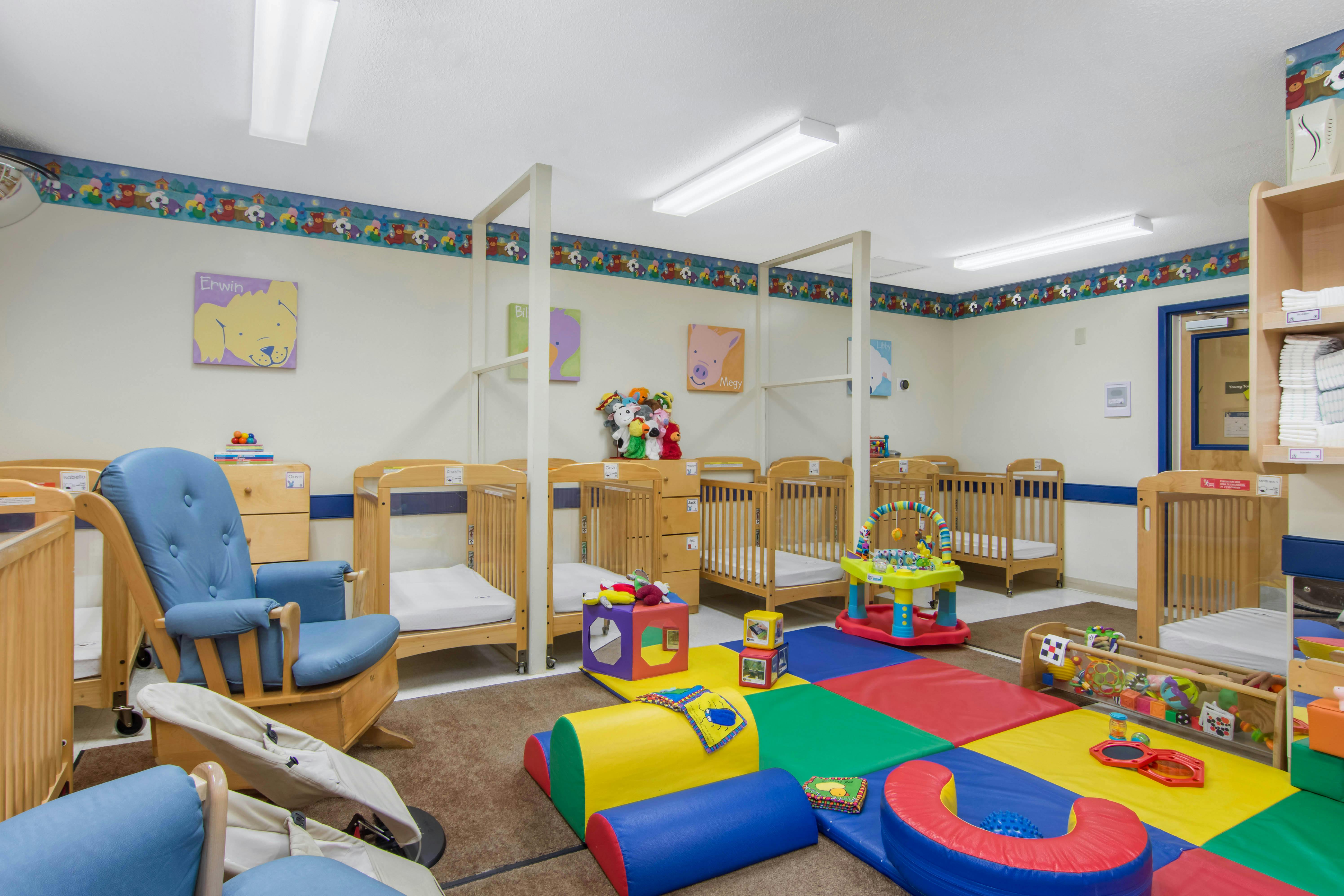 A room in a daycare with several cribs, a blue rocking chair, and colorful playmats