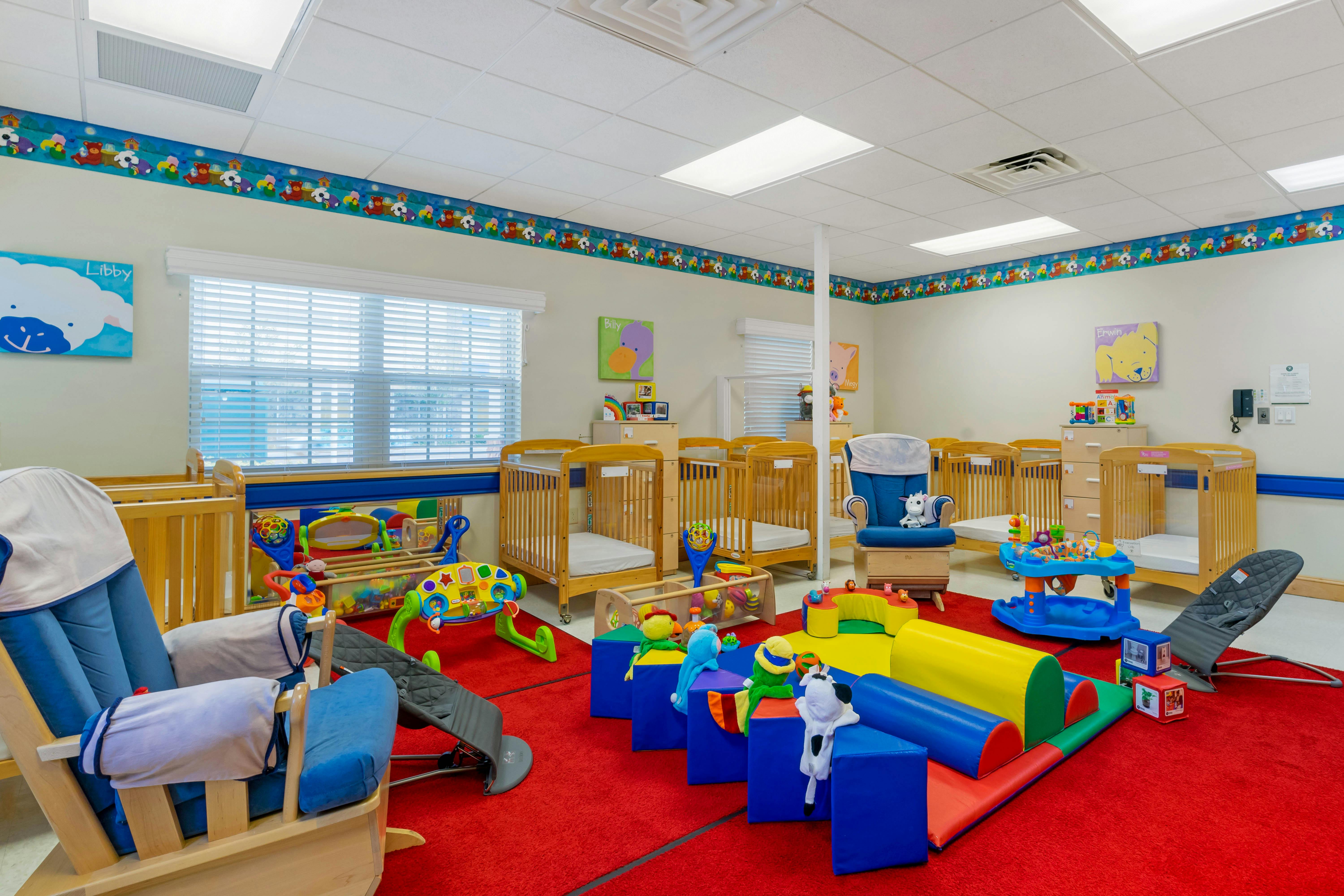 A colorful childcare classroom with multiple cribs, a blue rocking chair, and play equipment