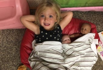 Child lying on a red cushion with two dolls wrapped in a blanket